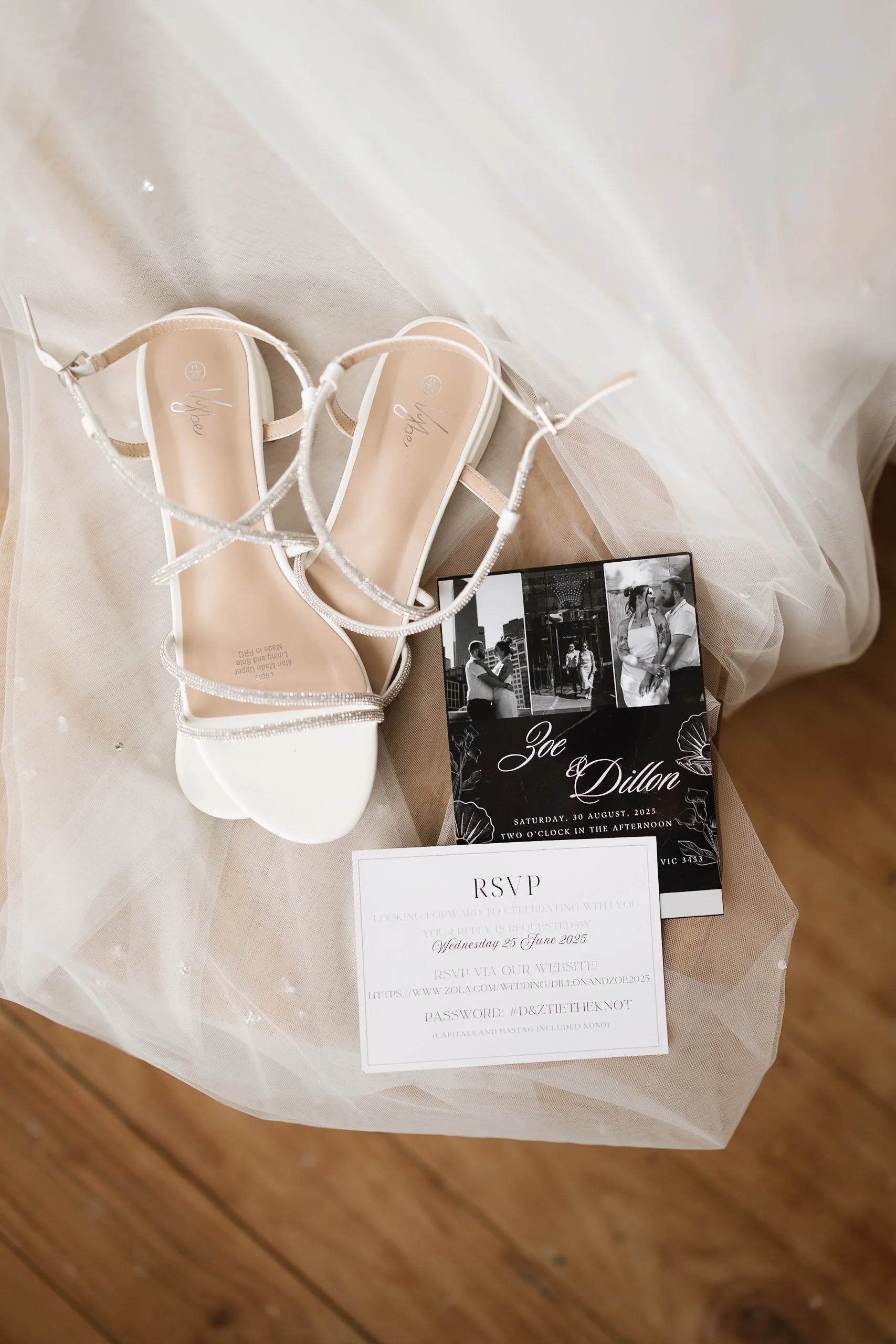A pair of white open-toe sandals with ankle straps sitting on a sheer veil or fabric. A black and white photo booth strip showing a couple and a wedding invitation for Joe and Dillon with wedding details, and an RSVP card with a response deadline and