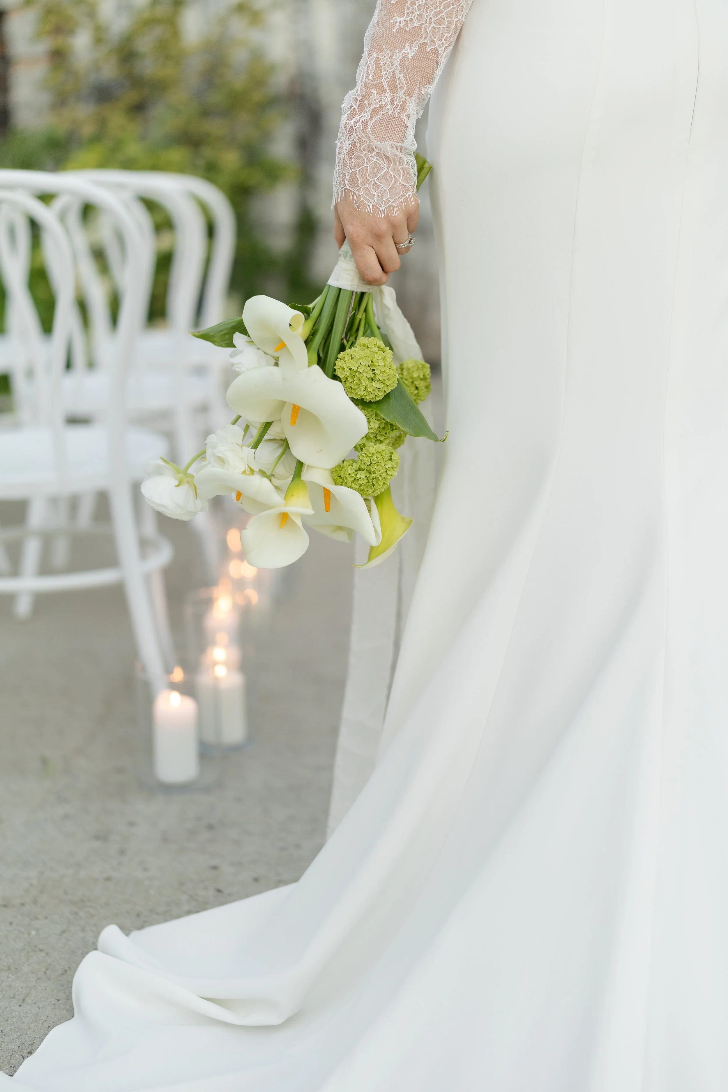 A bride in a white wedding dress holds a bouquet of white calla lilies, green hydrangeas, and other greenery. The background features white chairs and candles, indicating an outdoor wedding setting.