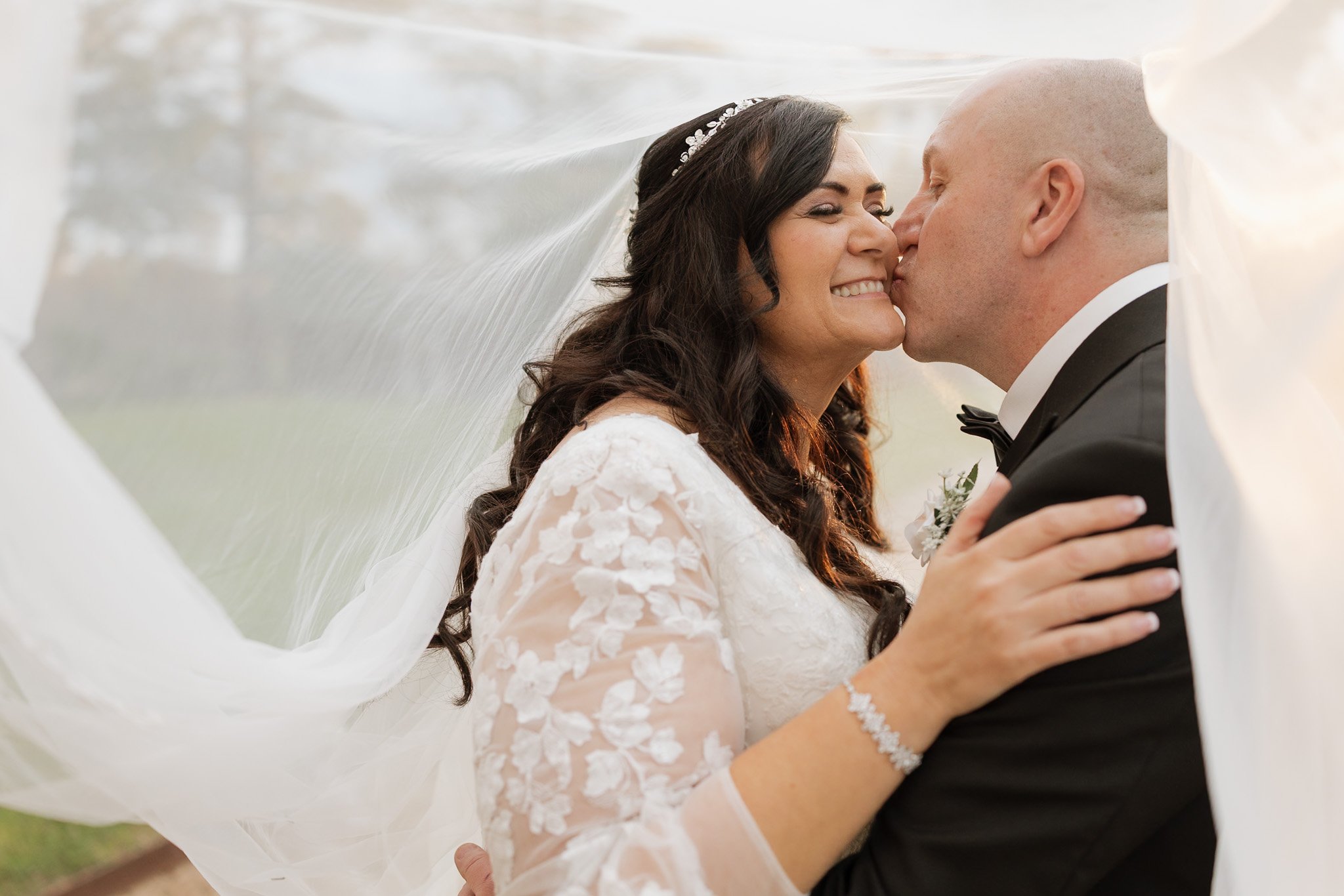 A bride and groom sharing a kiss outdoors beneath a wedding veil, with the bride wearing a lace wedding dress and the groom in a tuxedo.
