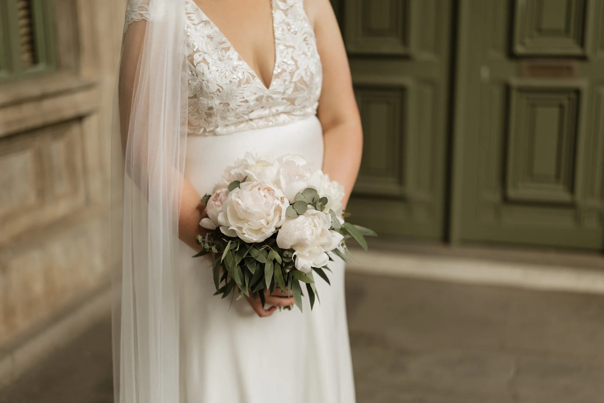 A bride in a white wedding dress holding a bouquet of white flowers and greenery, standing indoors with wooden panel walls.