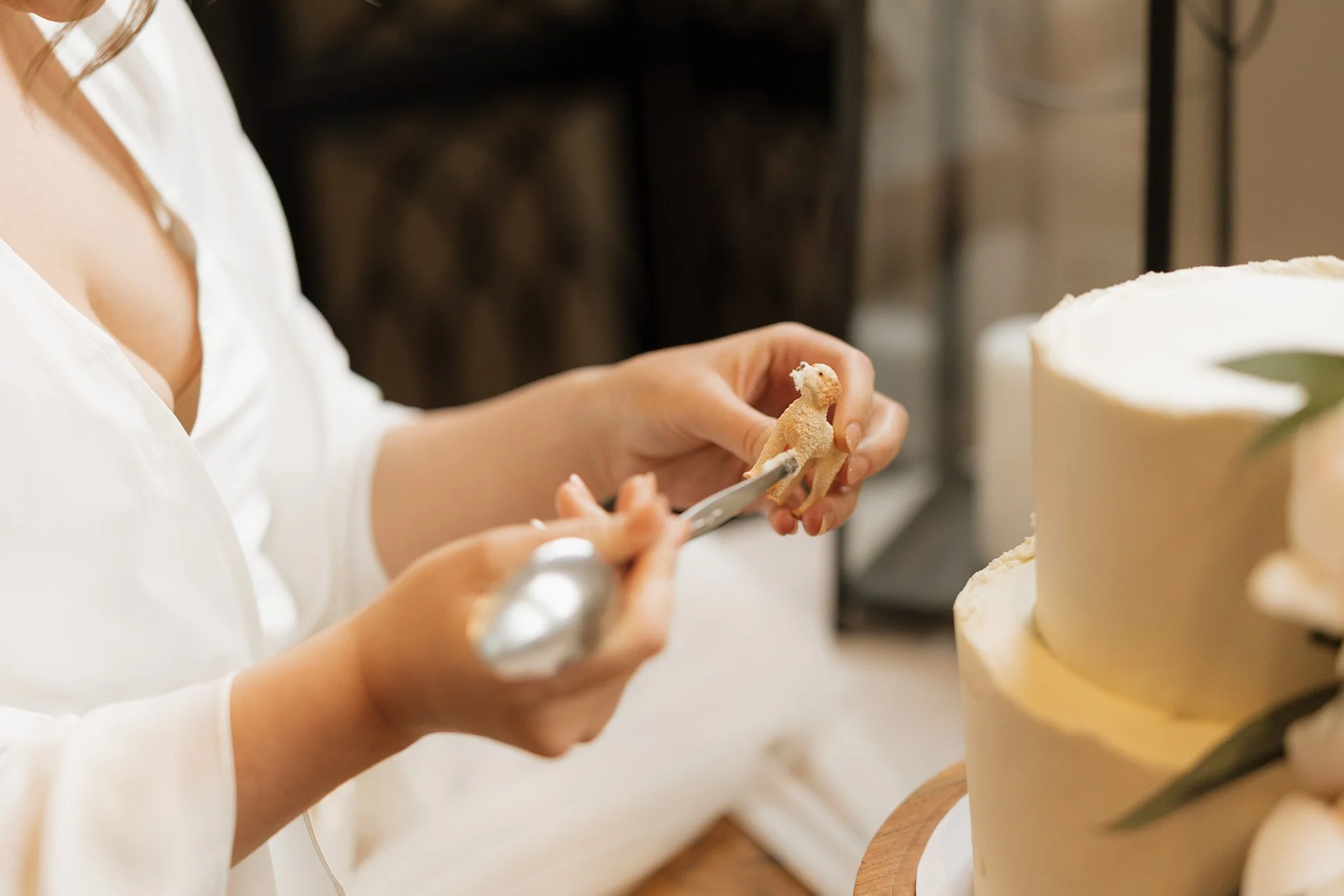 A person in a white shirt is carving a small, beige-colored figurine that resembles a dinosaur or dragon. There is a cake in the background with white icing and green decoration.