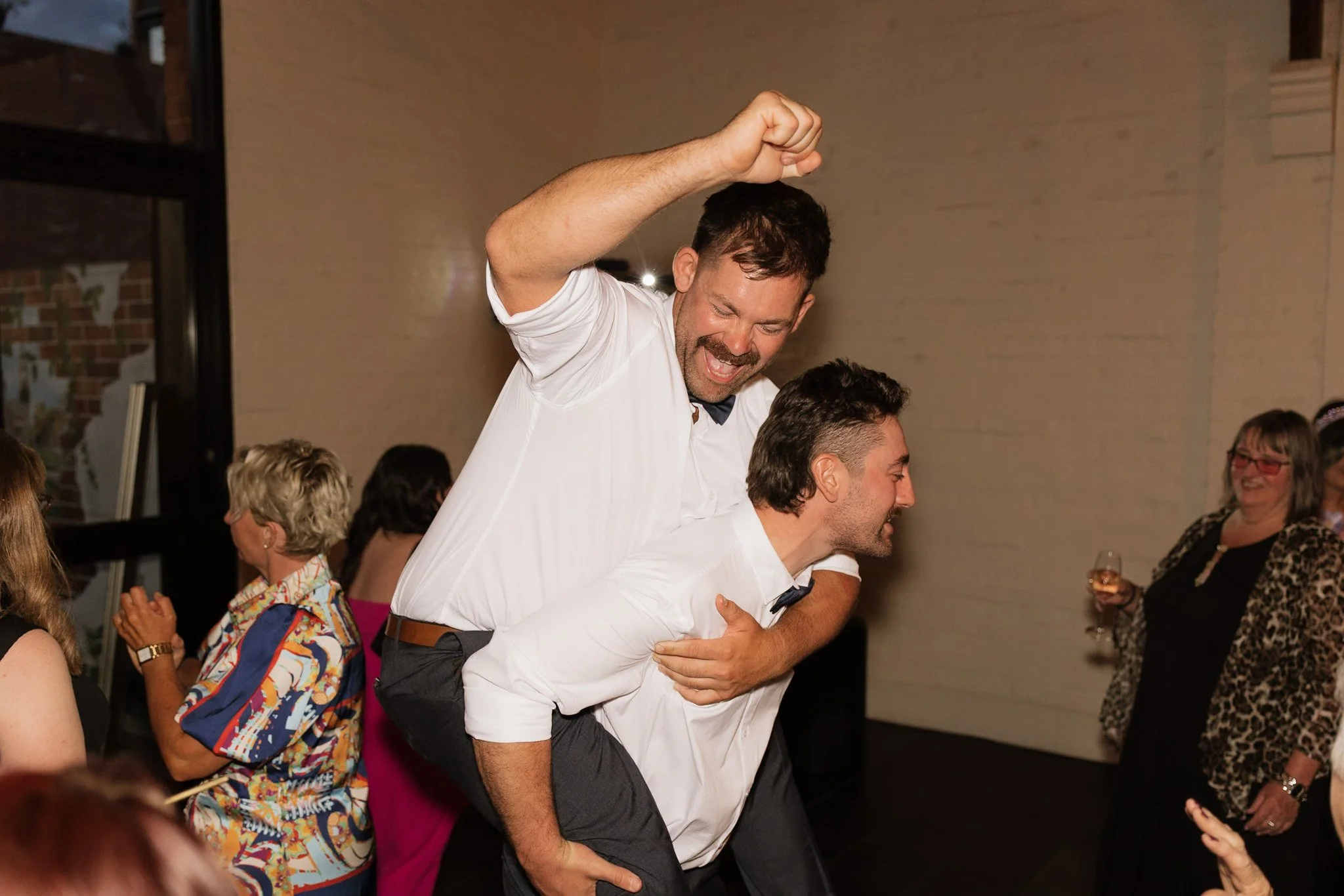 A man with dark hair and a beard, wearing a white shirt, is joyfully dancing on someone's back during a celebration event. The background features several women clapping and smiling, with one woman holding a wine glass. The setting appears to be indo