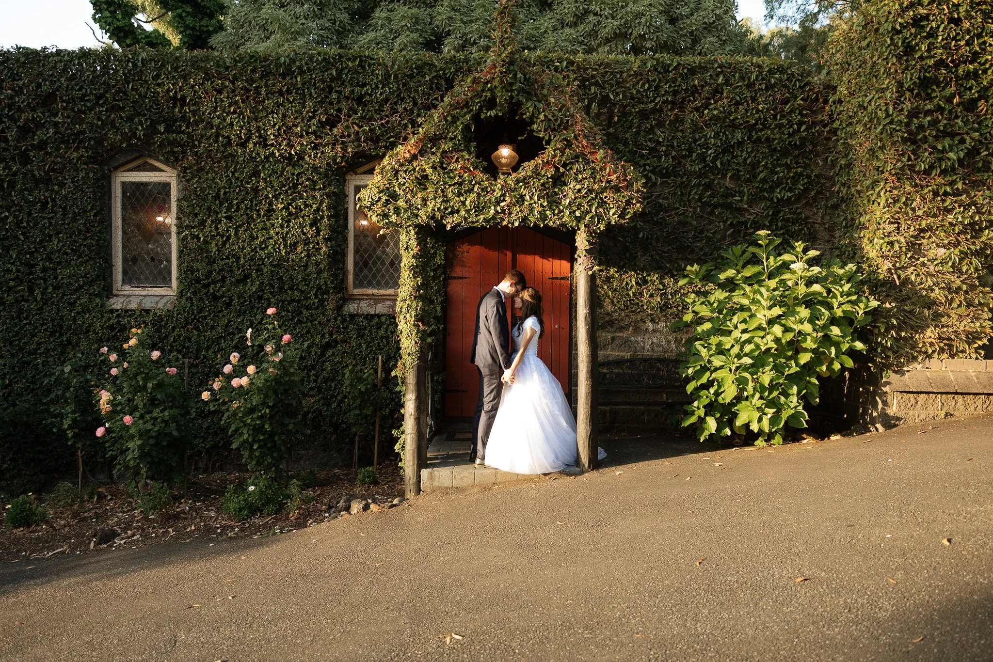 A bride and groom standing close together under a small wooden arch covered in greenery, sharing an intimate moment outside a building covered in ivy, with flowers and greenery surrounding the scene.