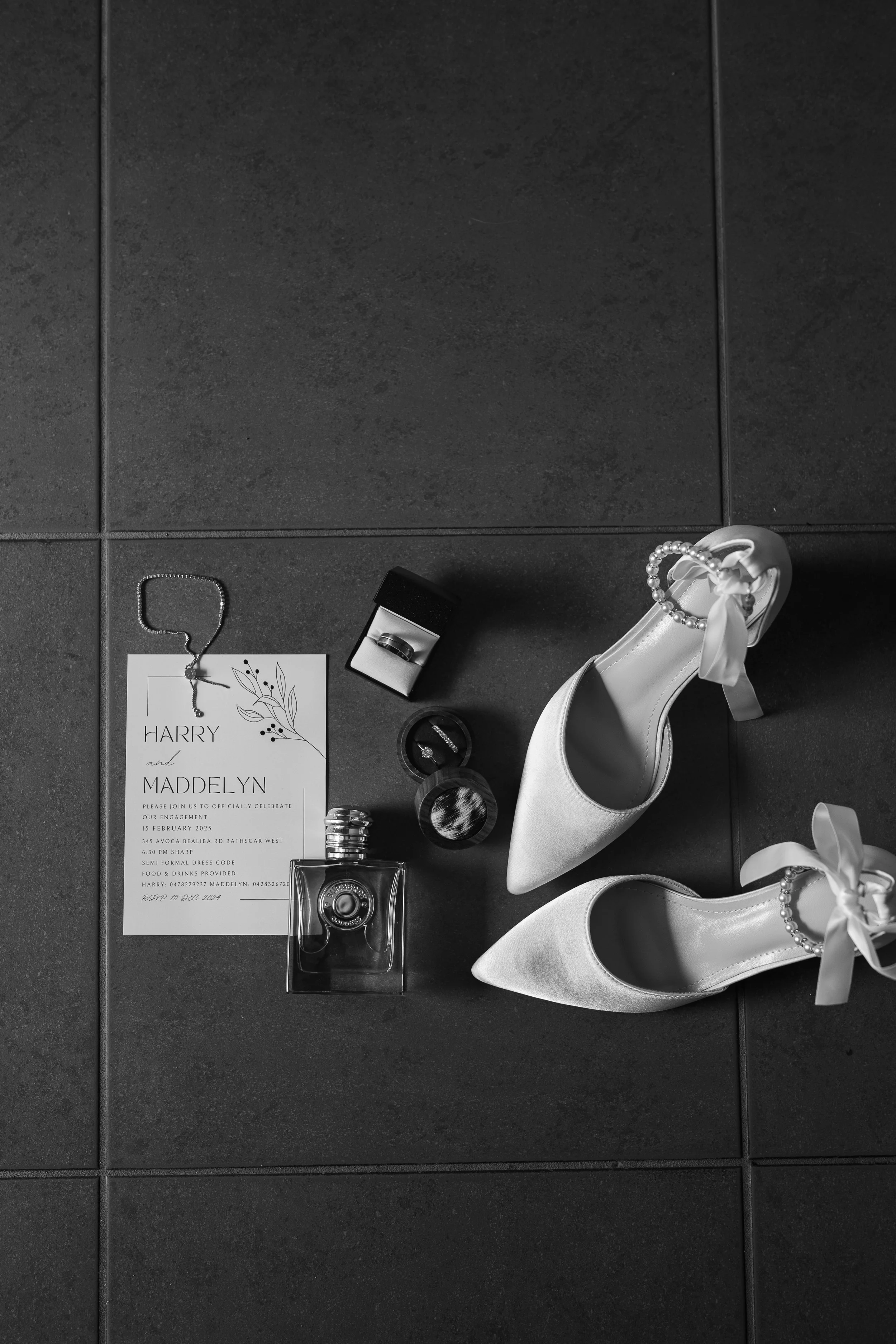 Black and white photo of a wedding invitation, pearl jewelry, a ring box, perfume bottle, and a pair of white high-heeled shoes with ribbon bows and pearl embellishments on a dark tiled floor.