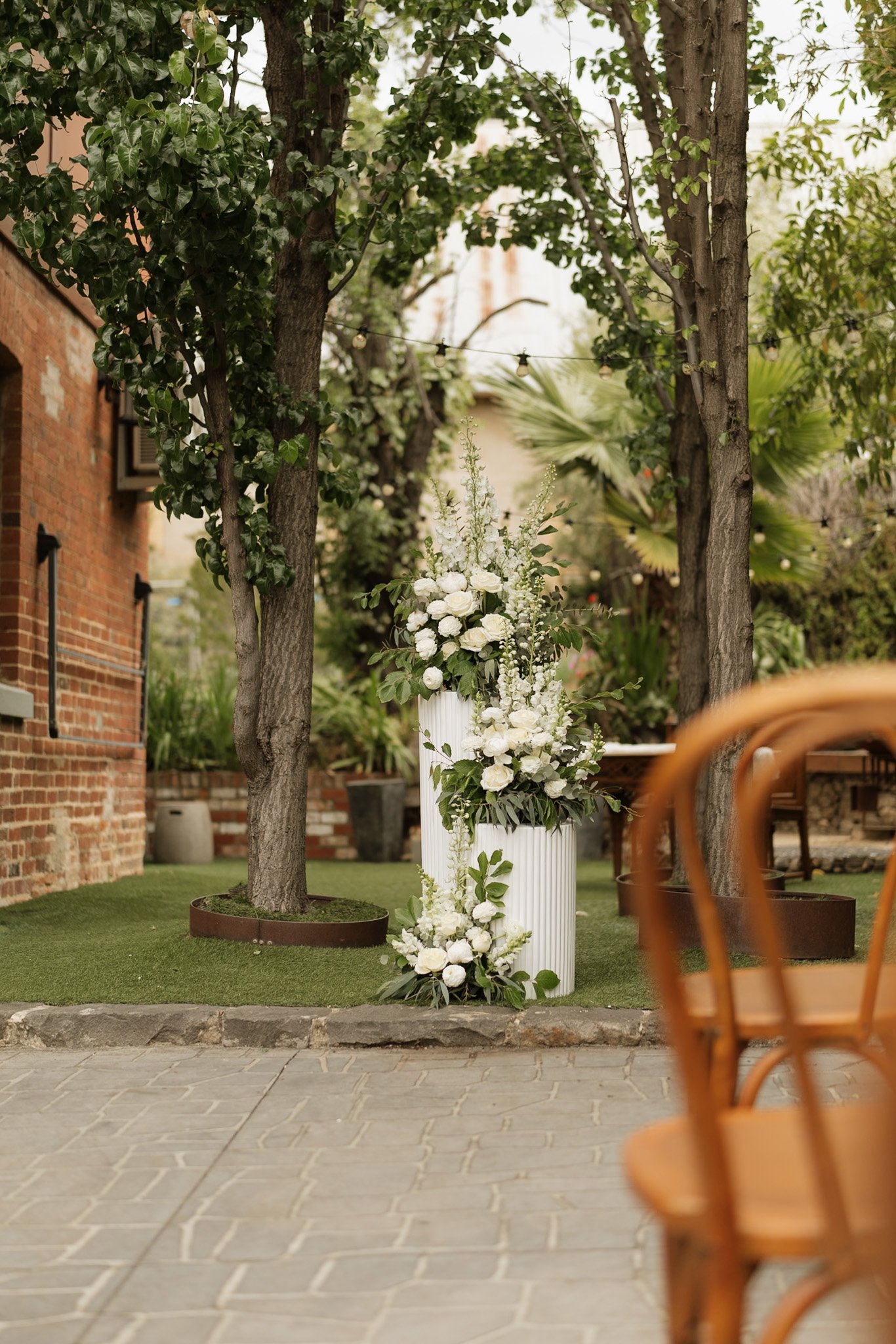Outdoor scene with trees, a flower arrangement in a white vase, and orange chairs in the foreground. There is a brick building on the left with a small window and potted plants, and string lights hang between the trees.