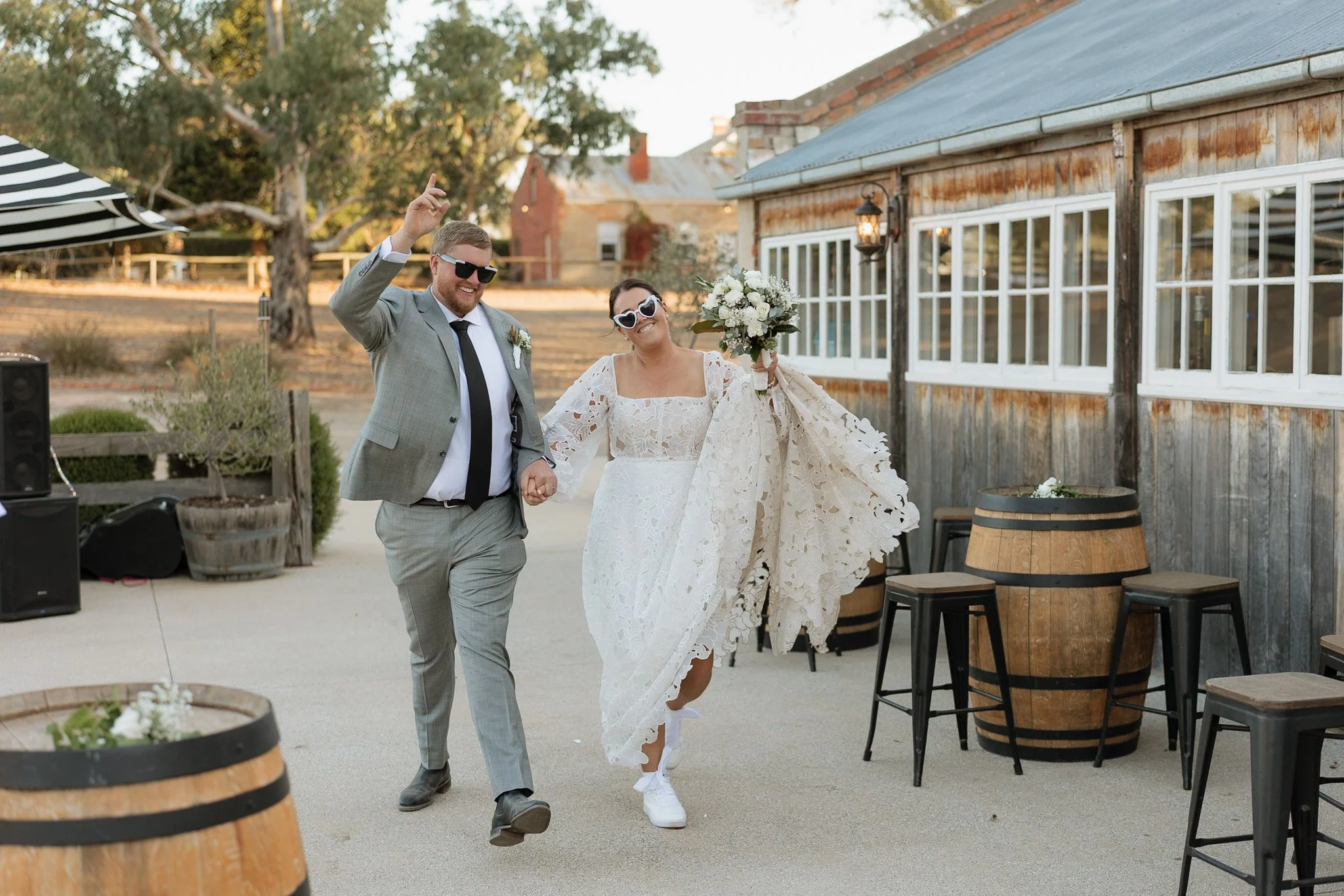A groom and bride holding hands and smiling, dancing outdoors at a wedding reception, wearing sunglasses and wedding attire.