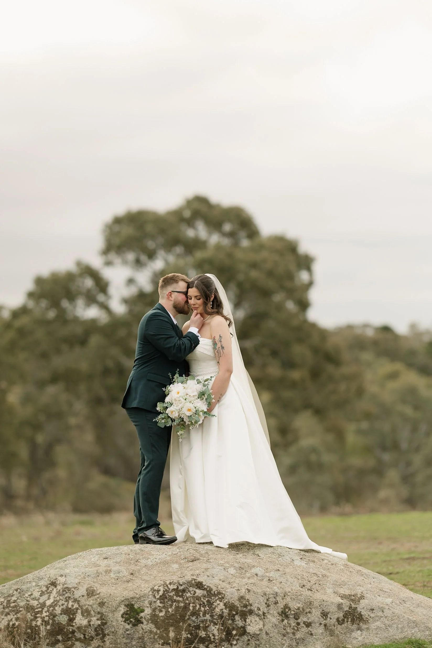 A bride and groom stand close together on top of a large rock outdoors, with trees and a cloudy sky in the background. The groom is dressed in a dark suit and glasses, leaning in to whisper or kiss the bride. The bride is wearing a white wedding gown