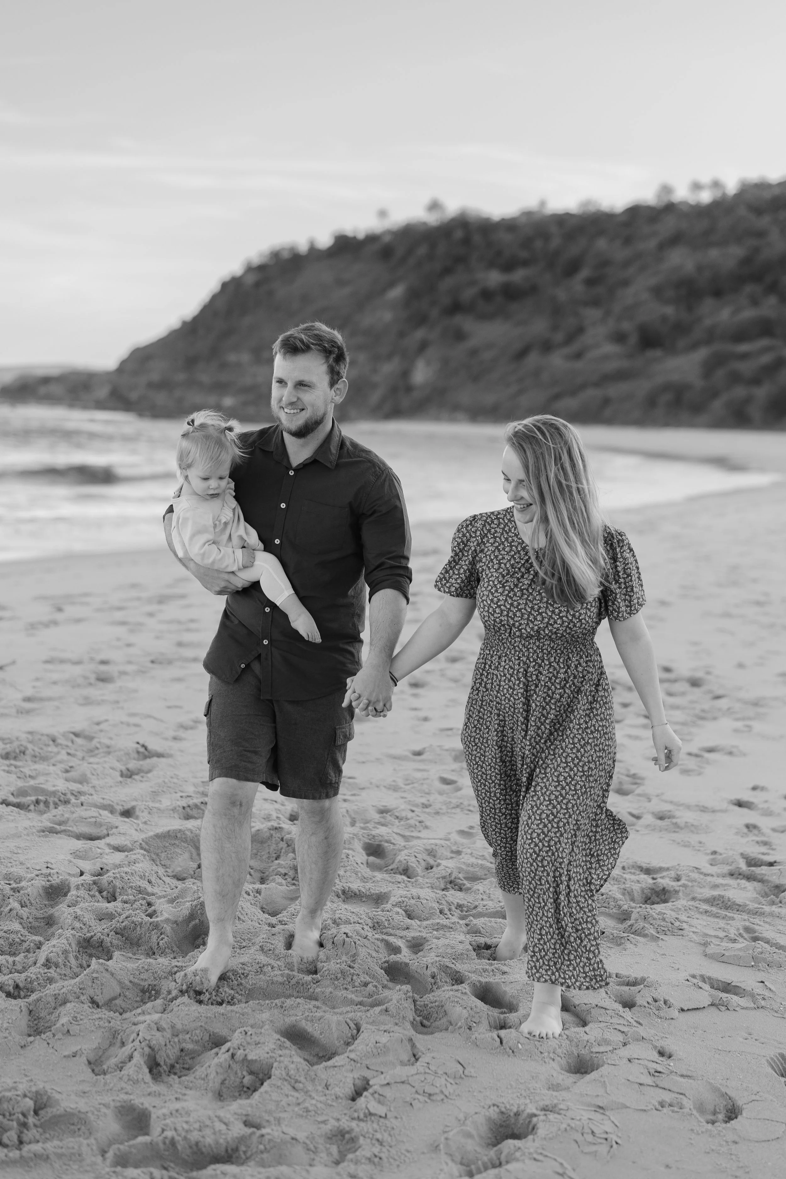 A happy family walking barefoot on the beach, holding hands, with a man carrying a young girl, and a woman smiling.