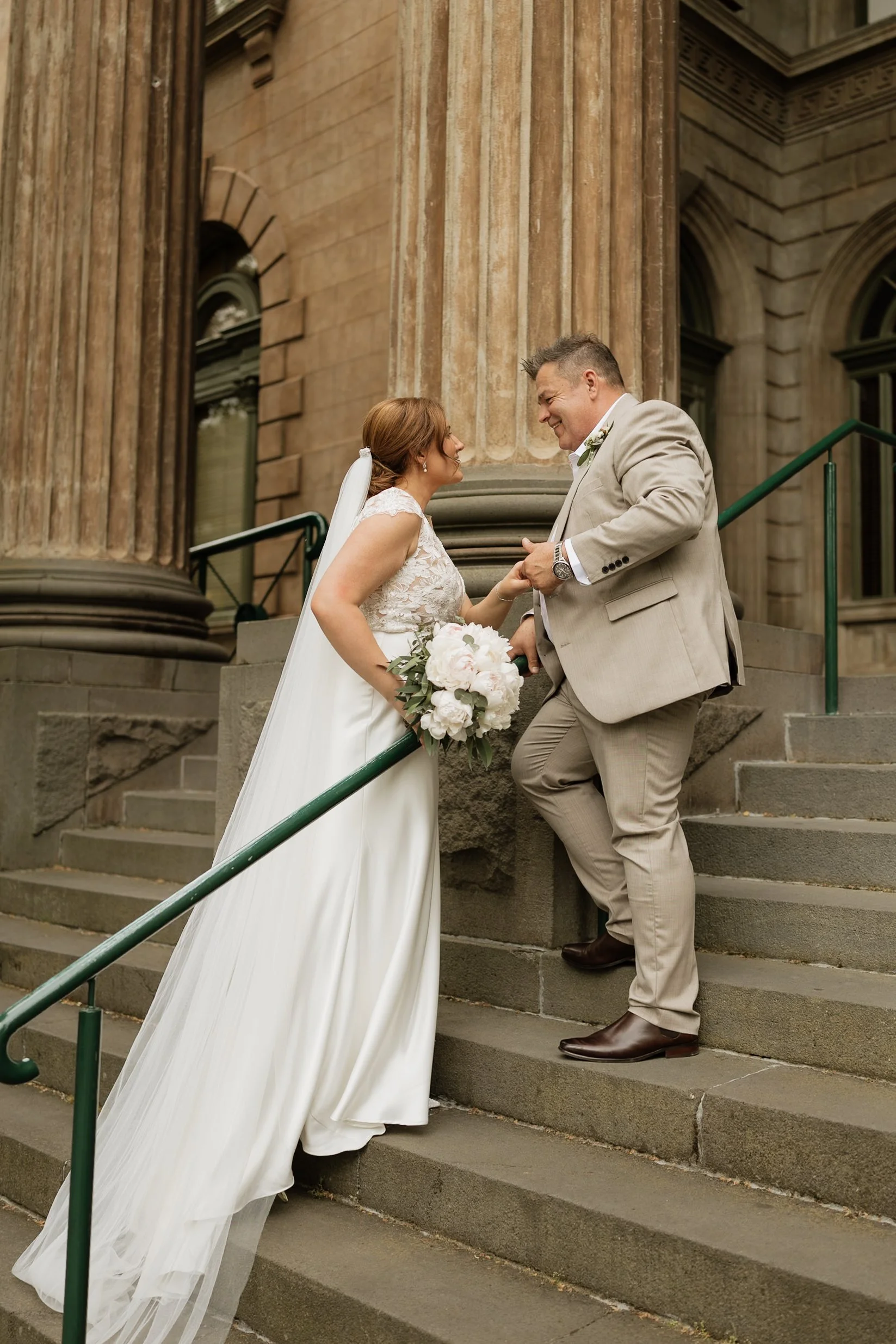 A bride and groom standing on the steps of a historic building, smiling and looking at each other, holding hands, with the bride holding a bouquet of white flowers. The bride is in a white lace gown with a veil, and the groom is in a beige suit with 