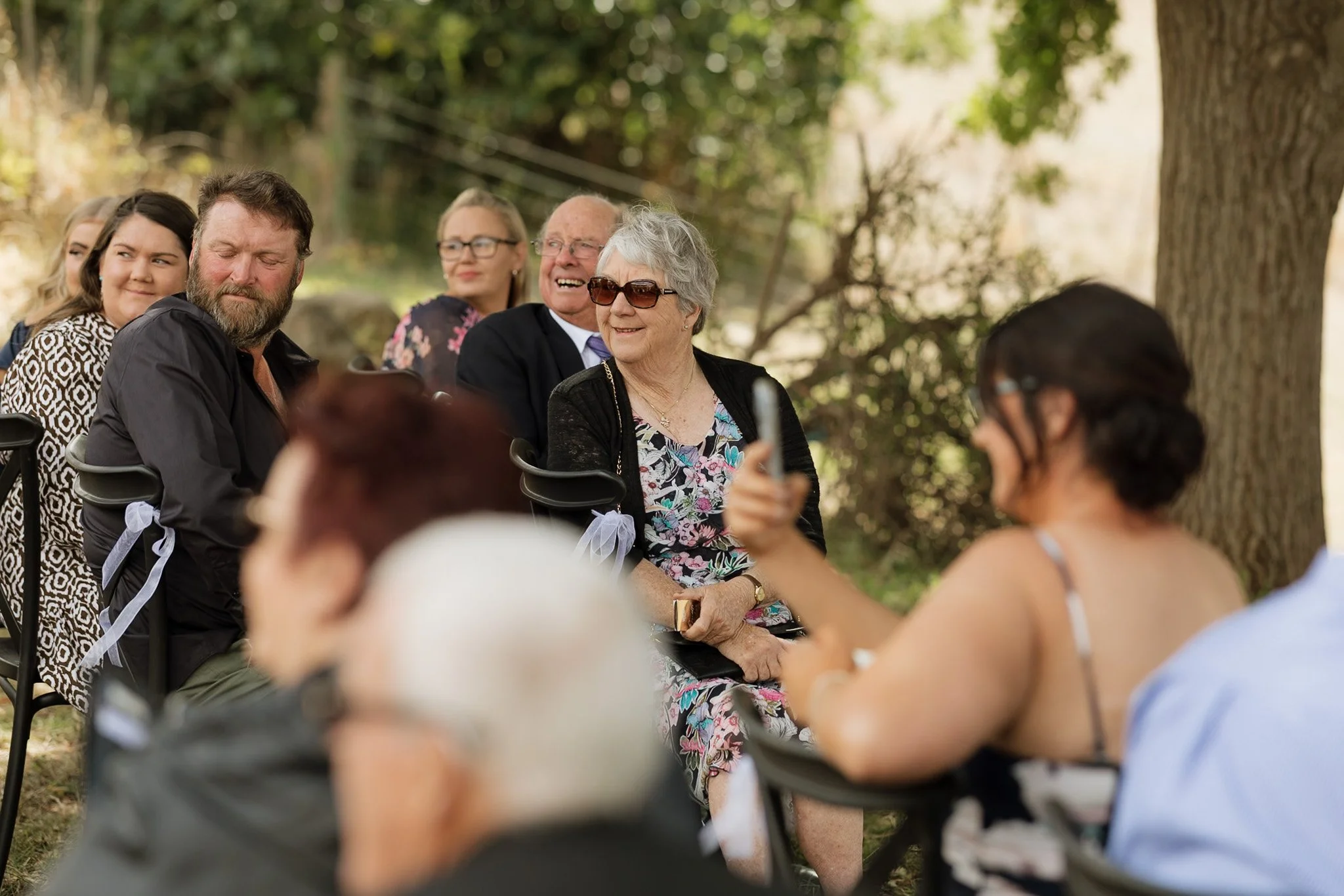 A diverse group of people sitting on chairs outdoors, attending a social event or gathering under trees, with some smiling and engaging with each other.