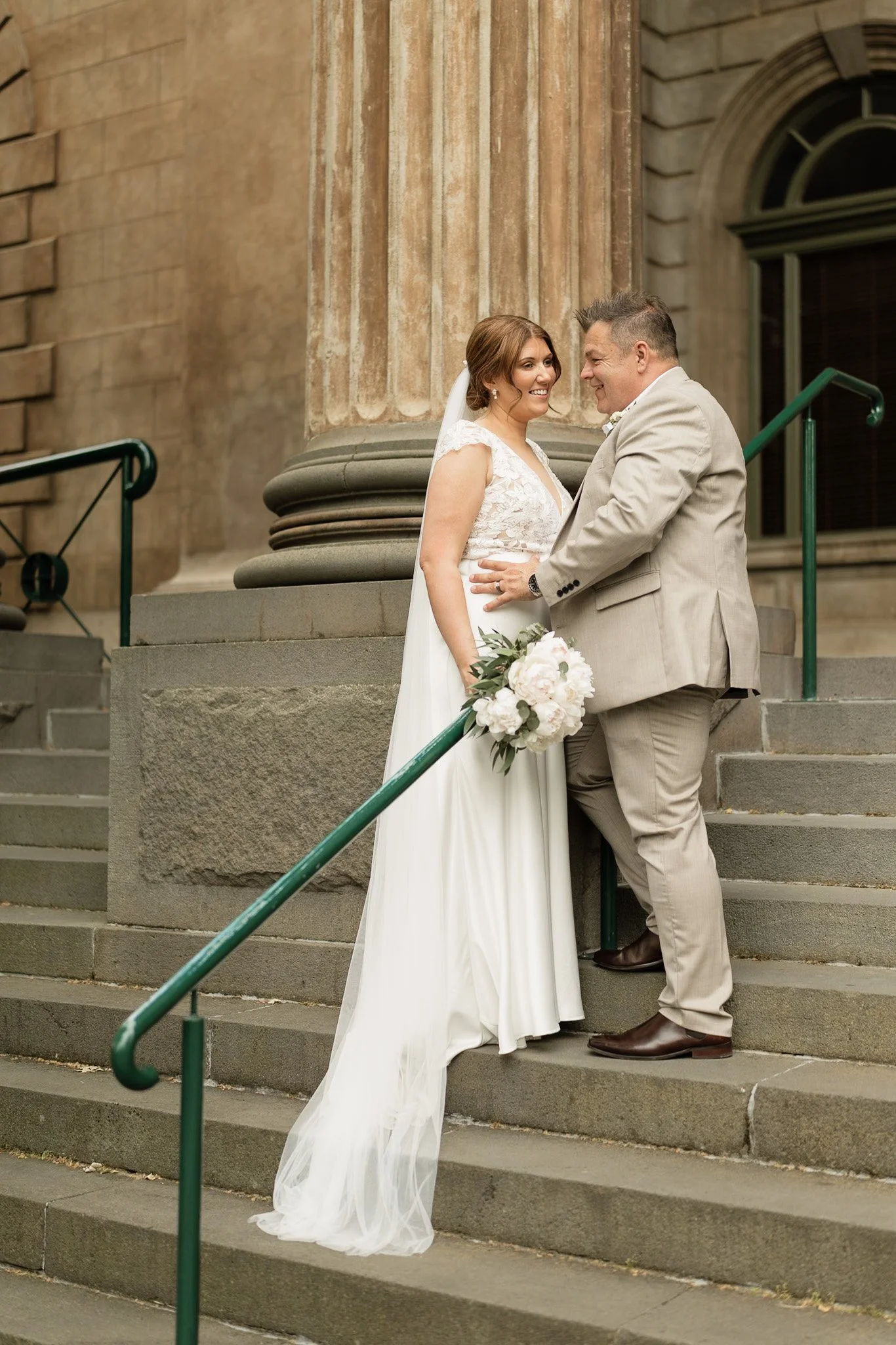 A bride and groom standing on stone steps in front of an old building with large columns and a window, smiling at each other. The bride is holding a bouquet of white flowers, wearing a white wedding dress with lace details and a veil. The groom is we