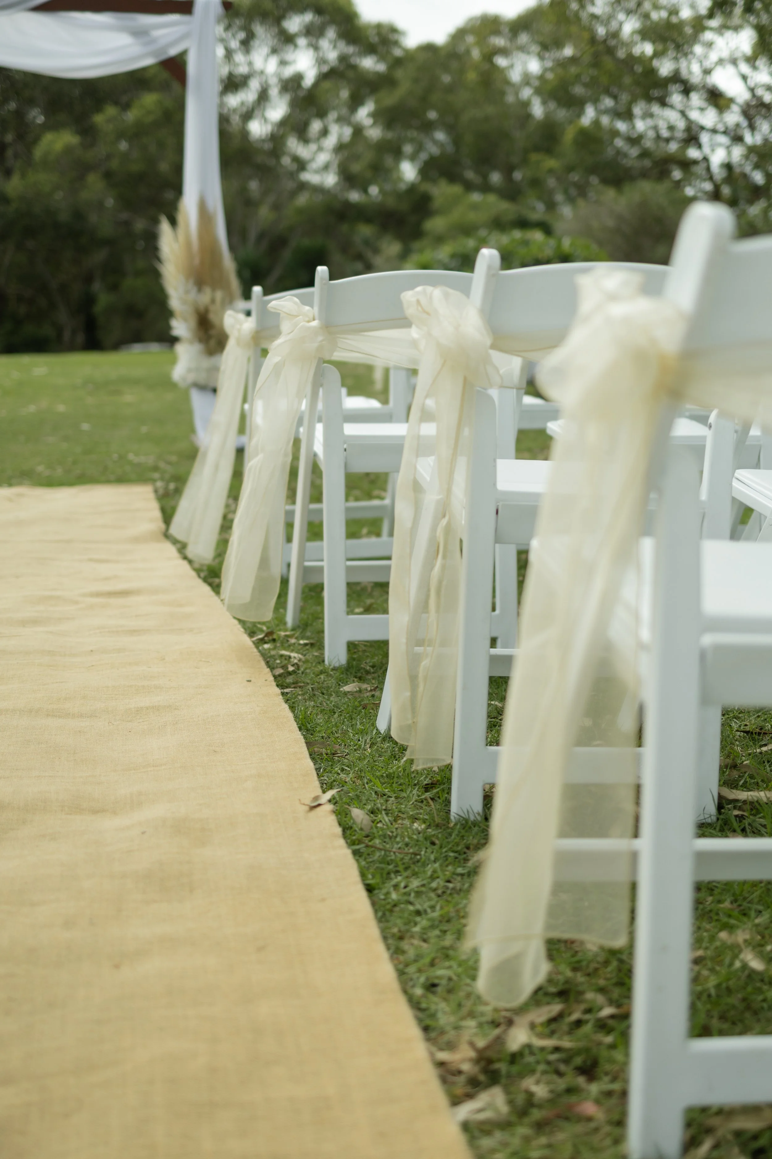 White chairs decorated with light yellow ribbons along a yellow aisle runner for an outdoor wedding ceremony in a grassy area with trees in the background.