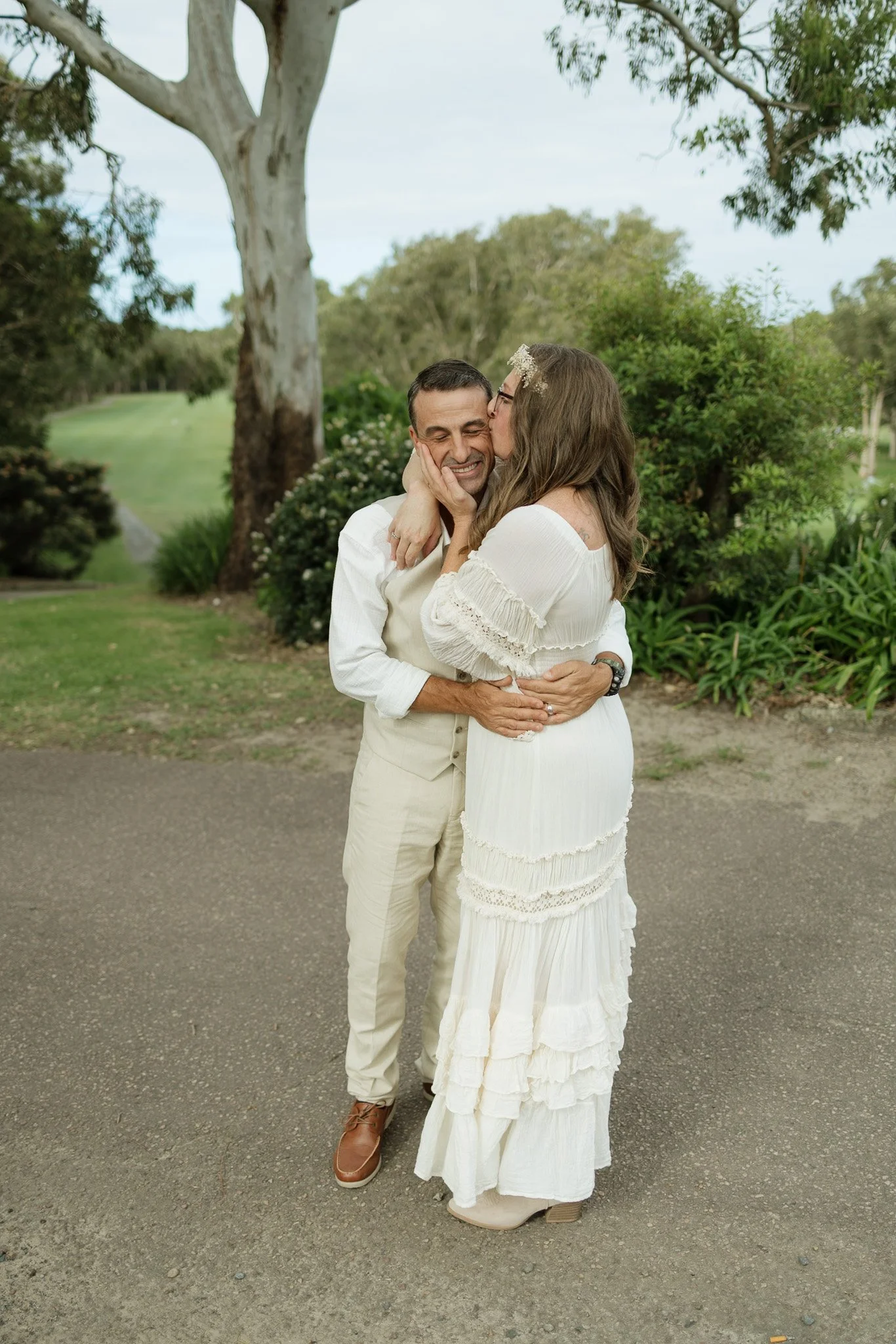 A couple appears to be close in the photo. The man has light skin, short dark hair, and is smiling while holding the woman. The woman has light skin, long brown hair, and is wearing glasses and a floral headband. She is sharing a kiss with the man's 