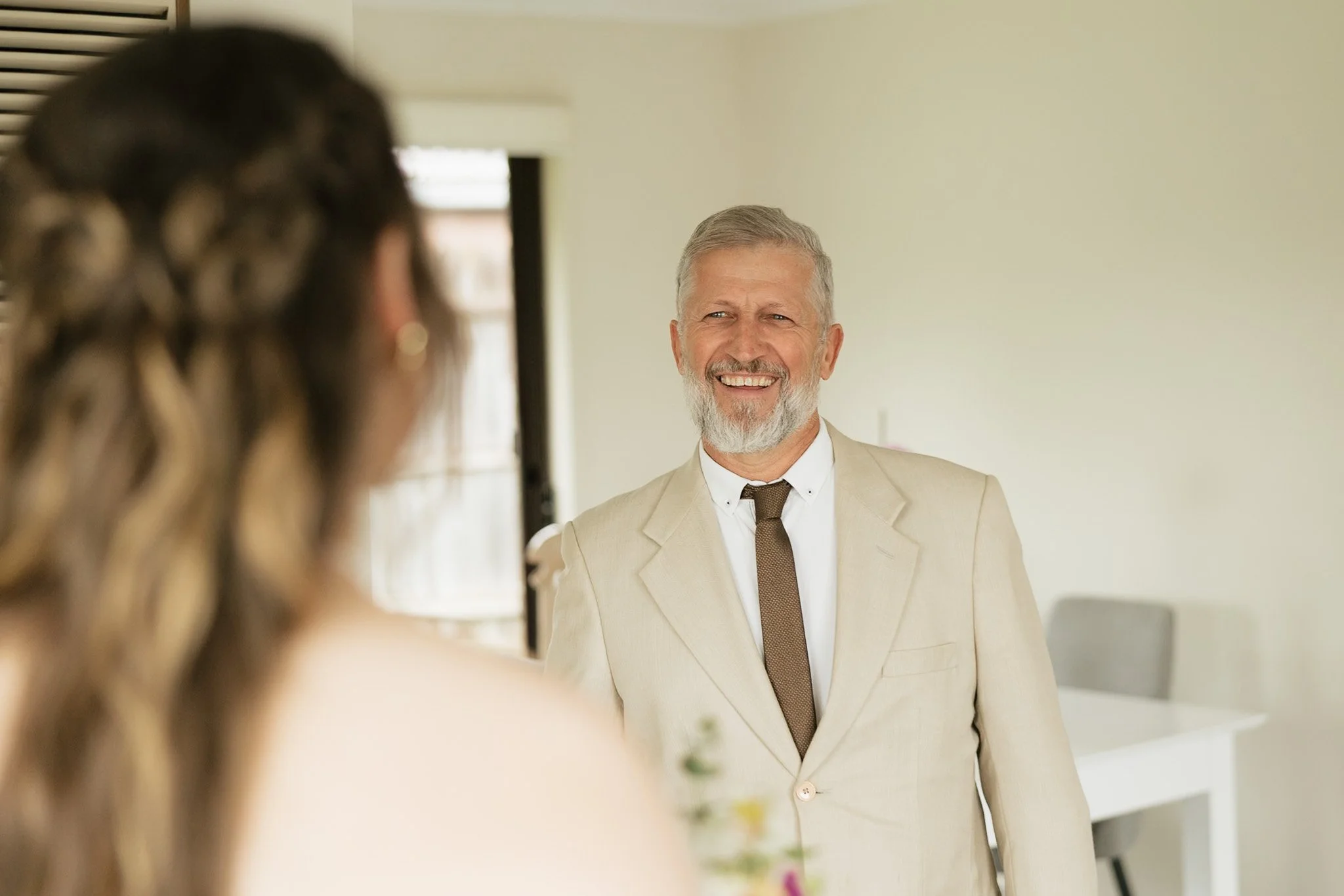 A man in a light beige suit with a white shirt and brown tie smiling at a woman with wavy hair, partially visible in the foreground, in a bright room.