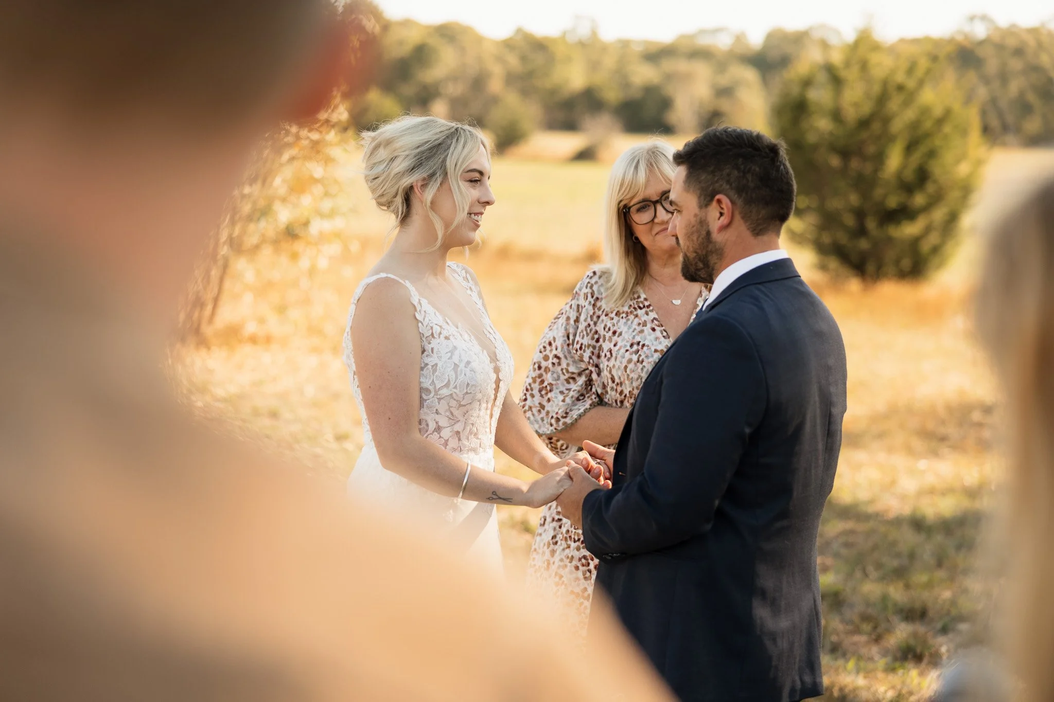 A wedding ceremony taking place outdoors on a sunny day, with a bride and groom holding hands and exchanging vows, an officiant standing between them, surrounded by trees and natural scenery.