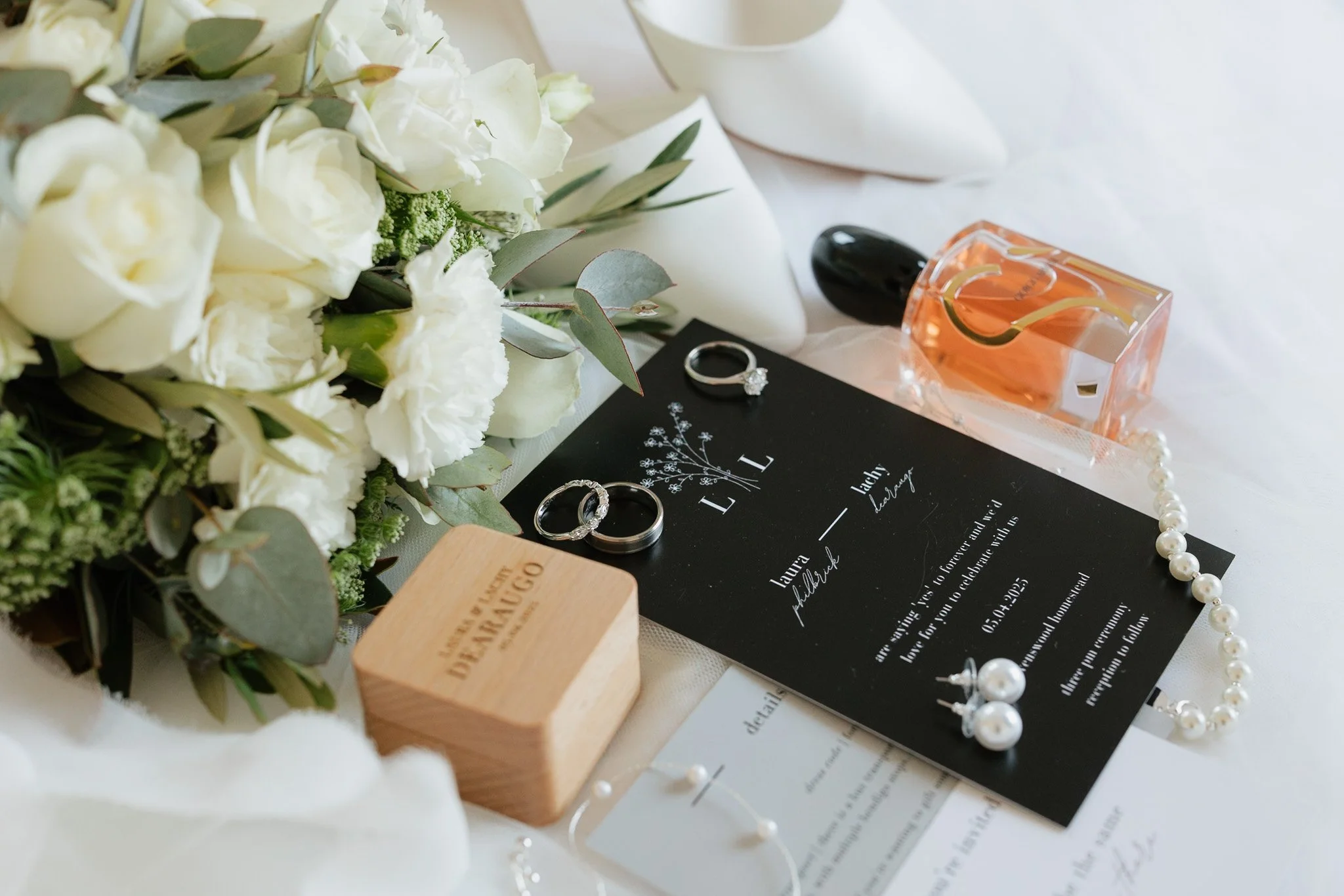 A bouquet of white flowers, a pair of silver engagement rings, a pearl necklace, a pink perfume bottle, and a wedding invitation on a white surface.
