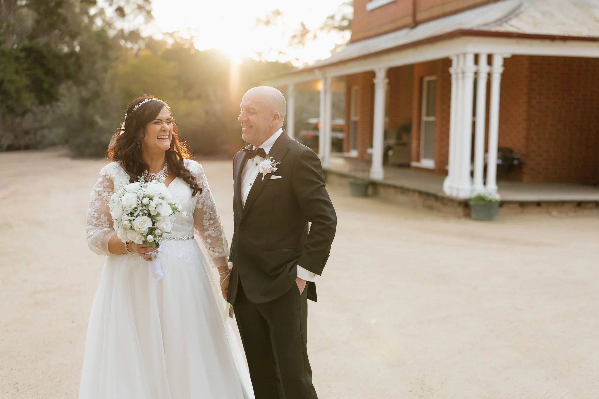 A bride and groom laughing together outdoors during sunset in front of a brick house with white columns. The bride is wearing a white wedding dress and holding a bouquet, while the groom is dressed in a black tuxedo with a bow tie.