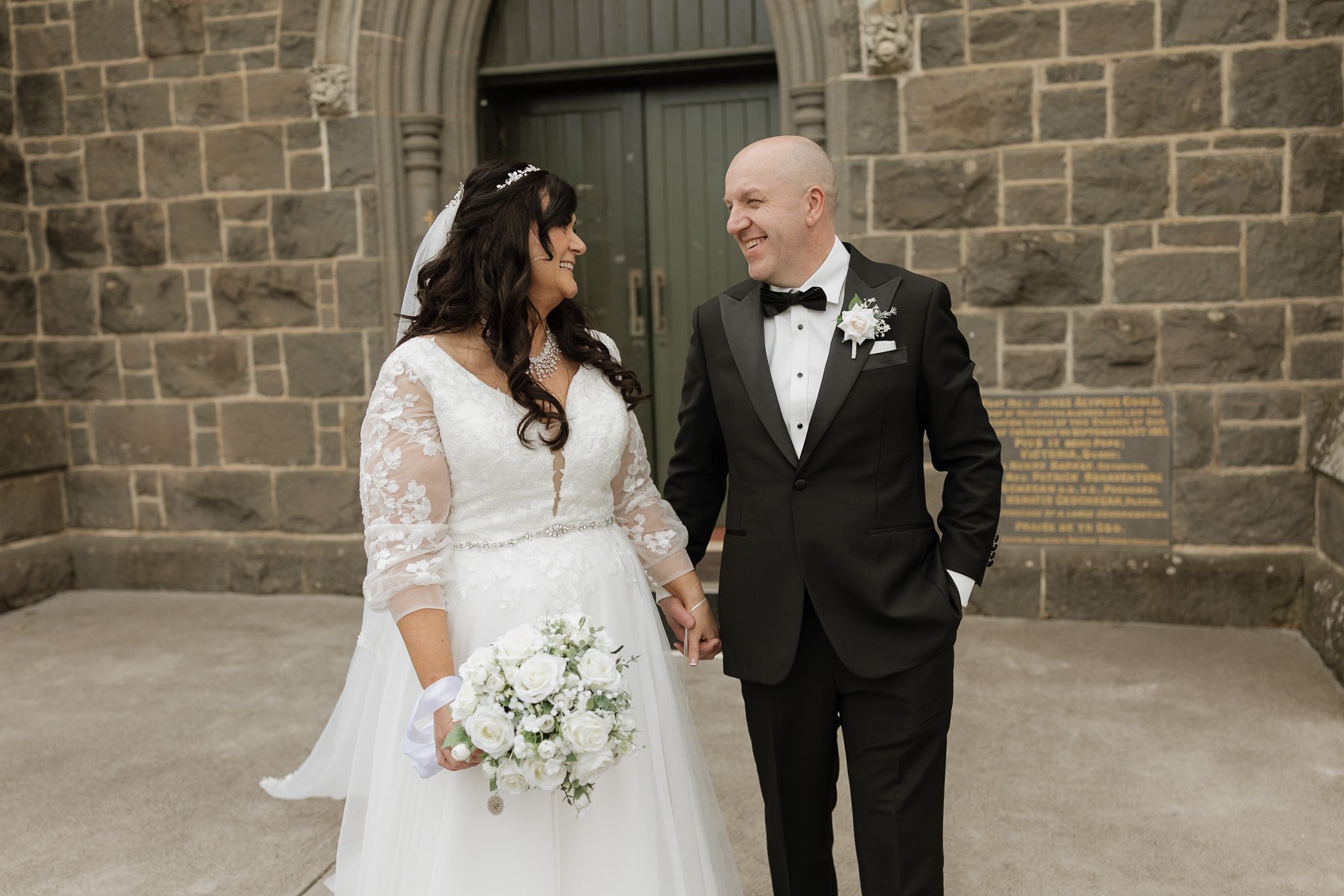 A bride and groom in wedding attire holding hands and smiling at each other outside a stone building.