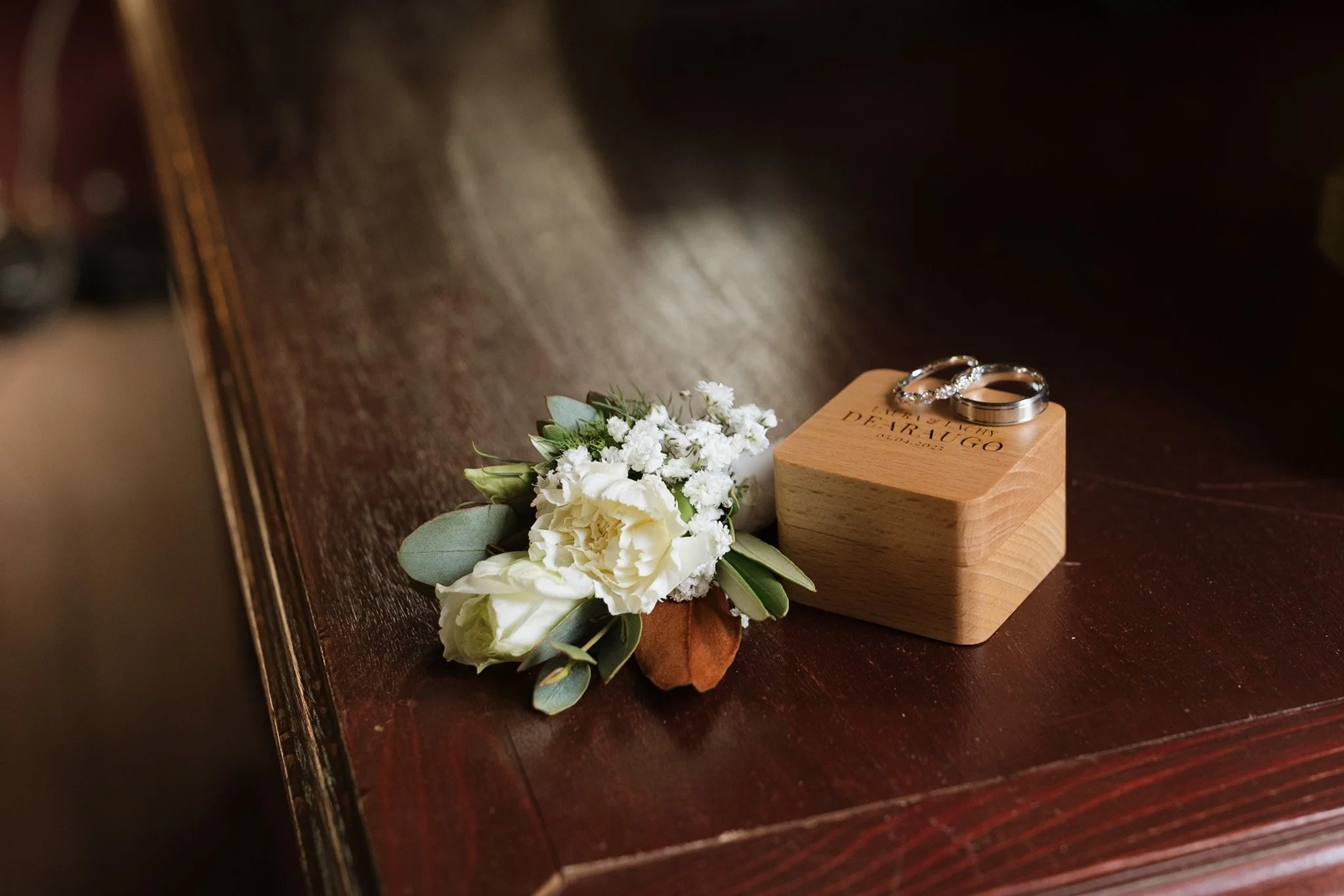 Two wedding rings, one with diamonds, placed on a small wooden box engraved with 'Wedding DeRUIGO 2023'. Next to the rings is a bouquet of white flowers with greenery, on a dark wooden surface.