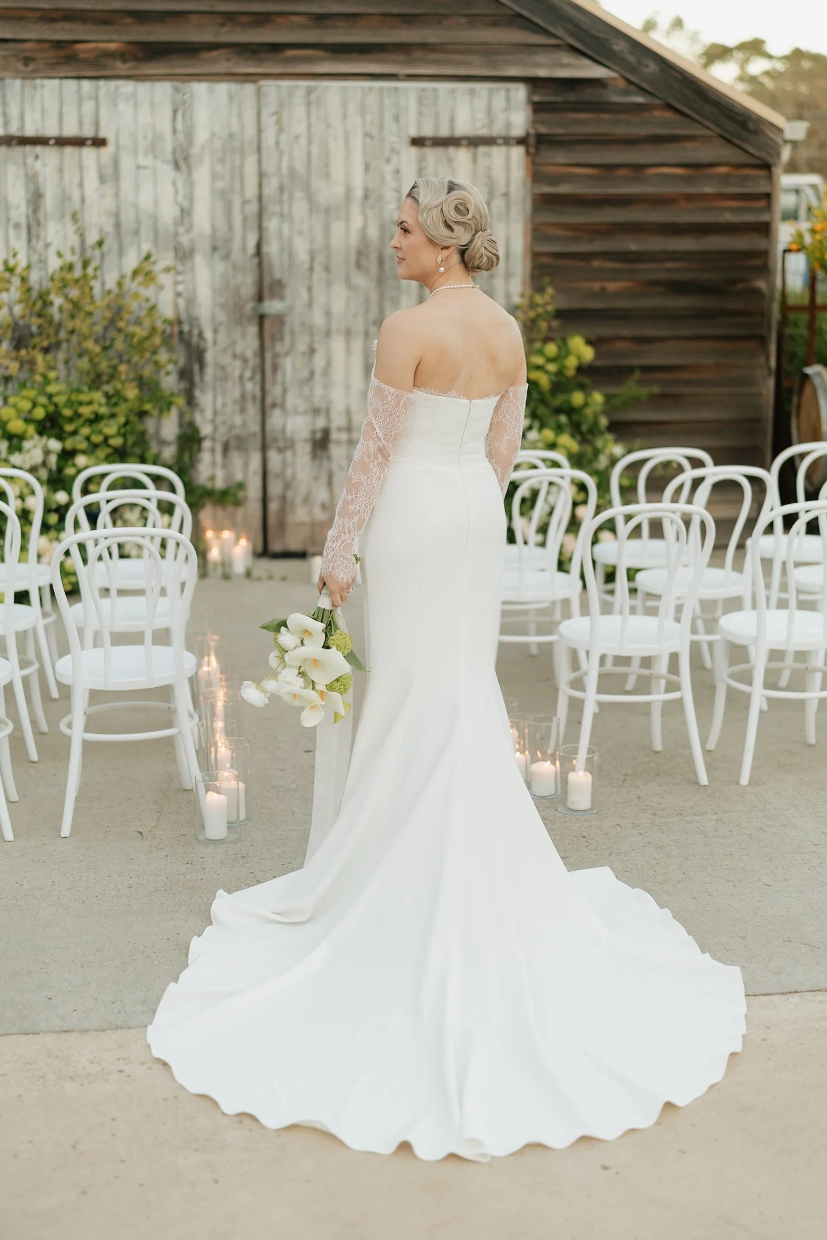 A bride in a white wedding dress with lace sleeves holding a bouquet of white calla lilies, standing outdoors with chairs and candles in the background.