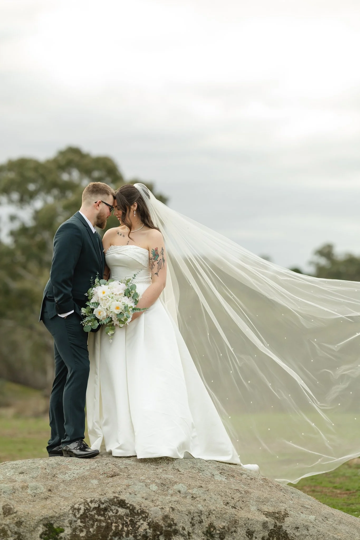 A bride and groom are standing close together on a rock outdoors, with their foreheads touching. The bride is wearing a white wedding dress with a long veil blowing in the wind, and holding a bouquet of white and pink flowers. The groom is dressed in a dark suit with glasses. Trees and a cloudy sky are in the background.