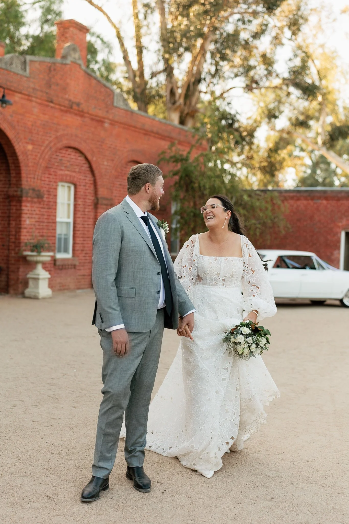 A newlywed couple outdoor, holding hands, laughing, dressed in wedding attire with a red brick building and trees in the background.