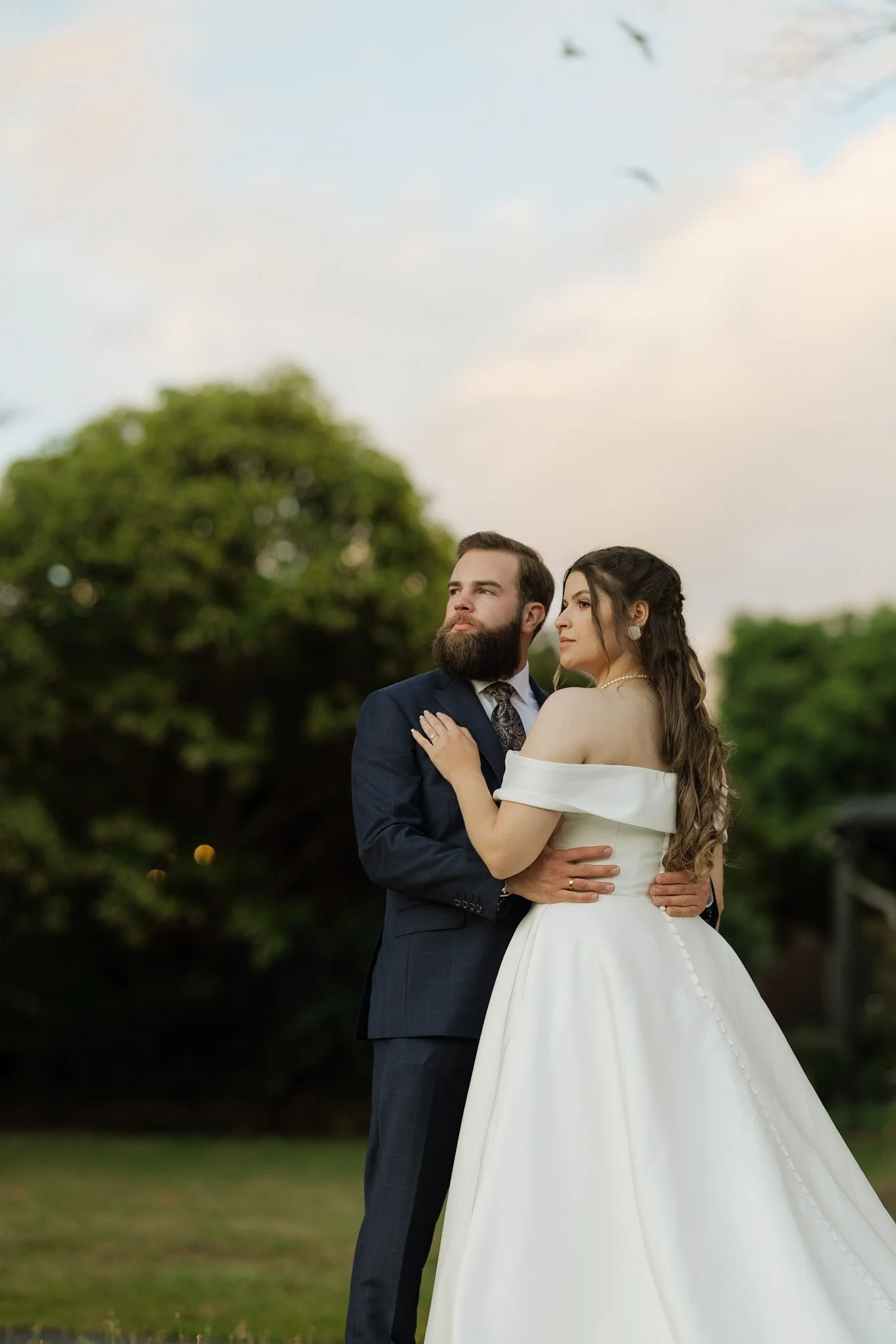 A couple dressed in elegant wedding attire standing outdoors, embracing each other with trees in the background and a partly cloudy sky overhead.
