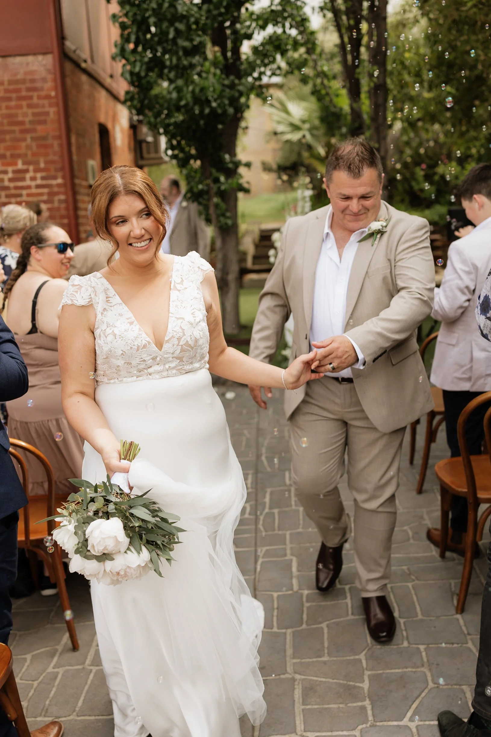 A bride in a white wedding dress holding a bouquet of flowers, smiling as she walks outdoors at a wedding celebration, with guests in background.