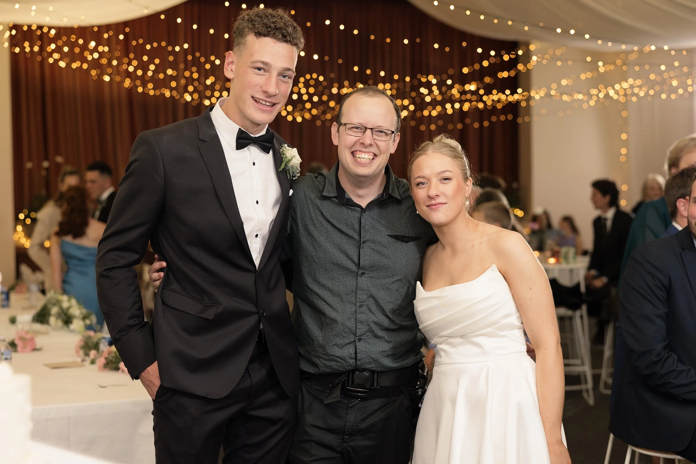 Three people smiling and posing for a photo at a wedding reception, with a decorated ceiling and tables in the background.