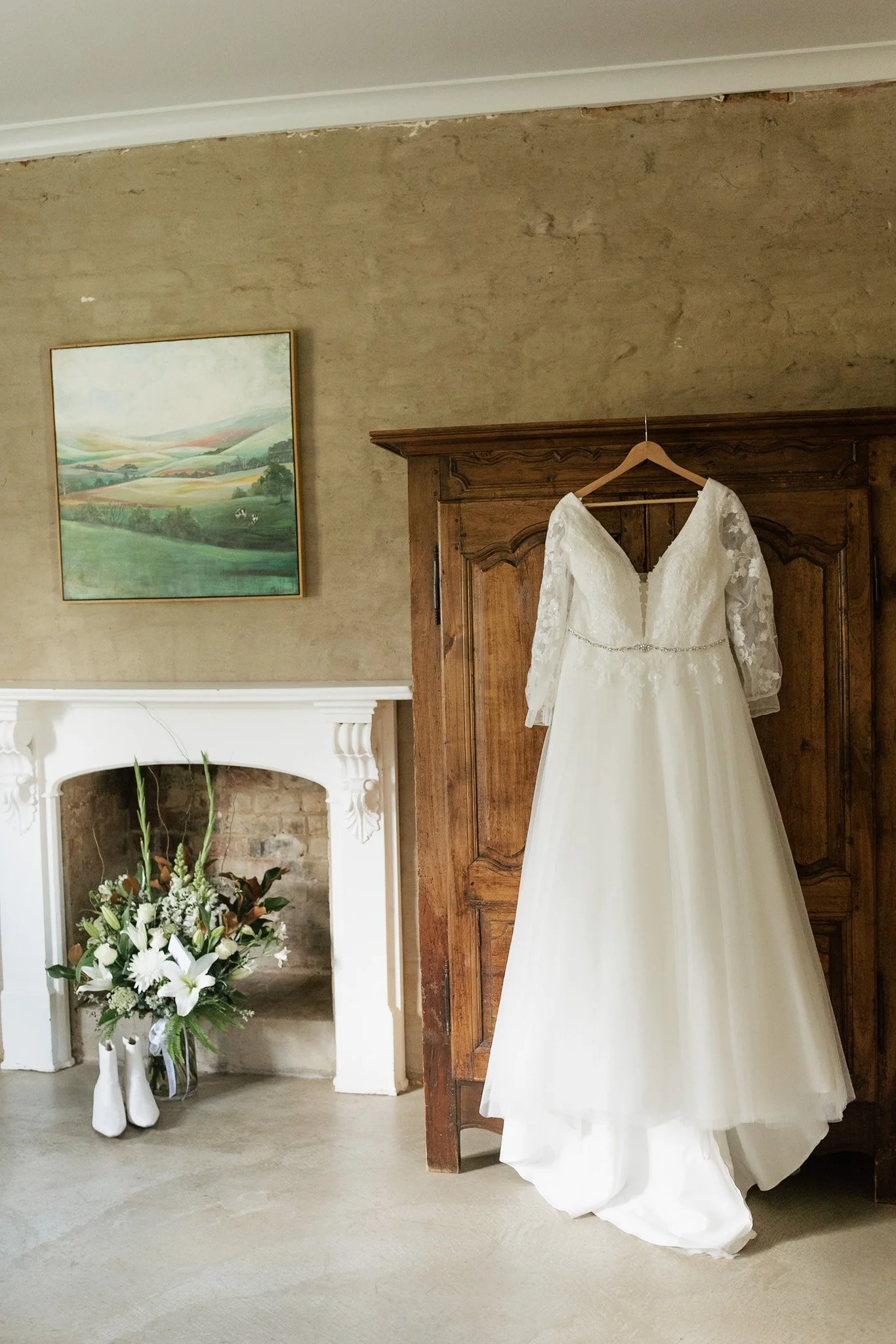 Wedding dress hanging on a wooden wardrobe in a room with a fireplace and floral arrangement.