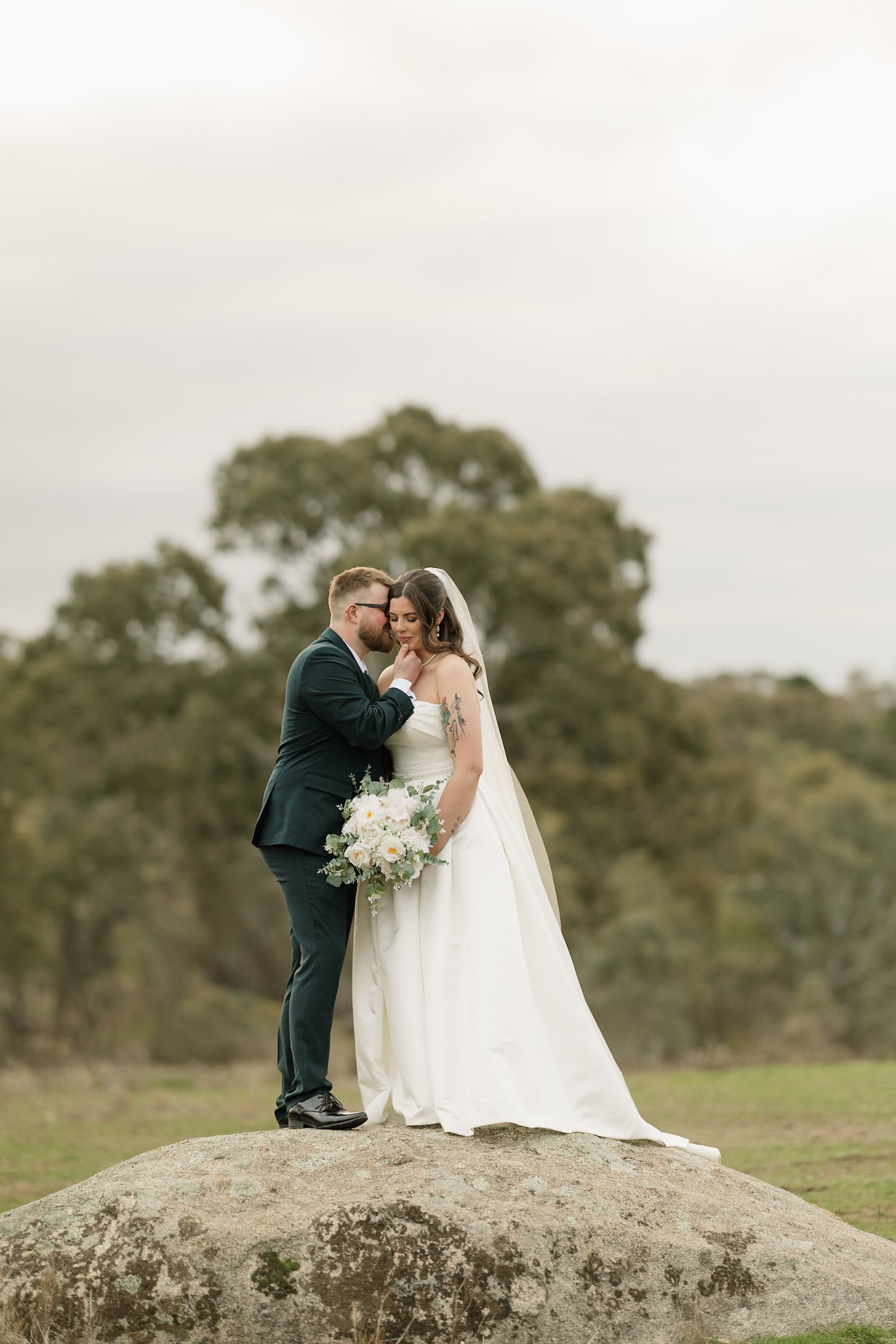 A bride and groom standing on a large rock outdoors, embracing each other. The bride is holding a bouquet of white flowers, and they are dressed in wedding attire. The background features trees and a cloudy sky.