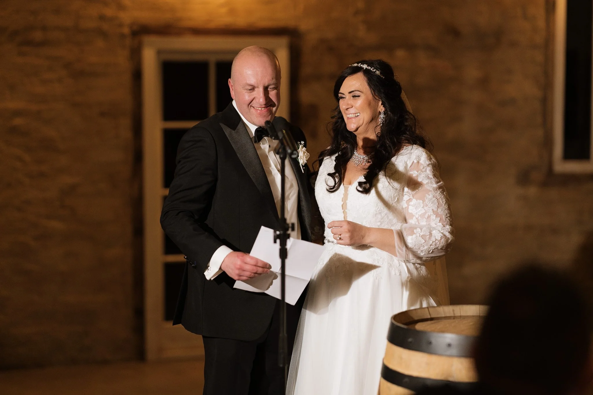 A bride and groom standing together during their wedding ceremony, smiling and reading vows in a rustic indoor setting.