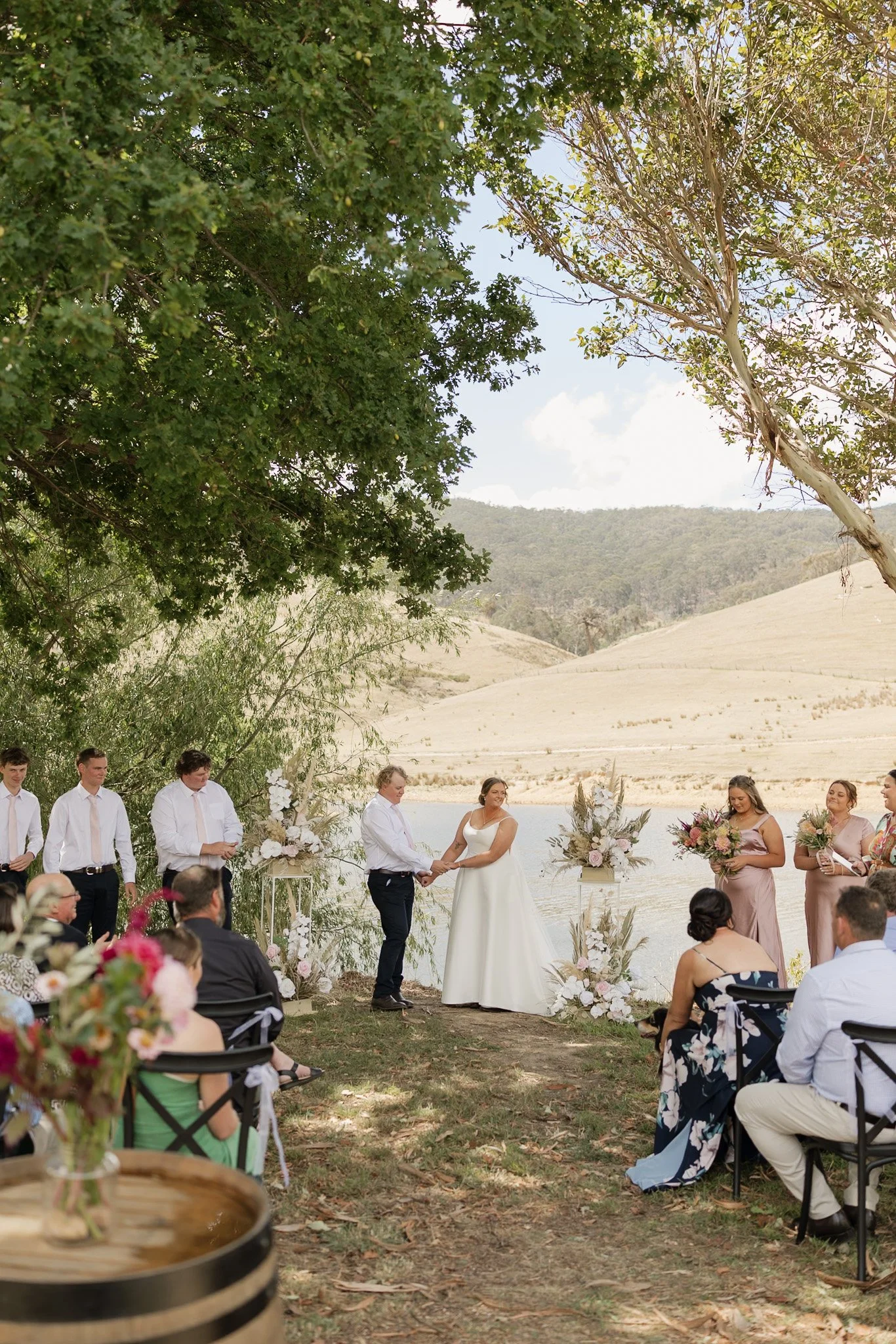 Outdoor wedding ceremony with bride and groom holding hands, surrounded by guests seated under trees near a lake and hilly landscape.