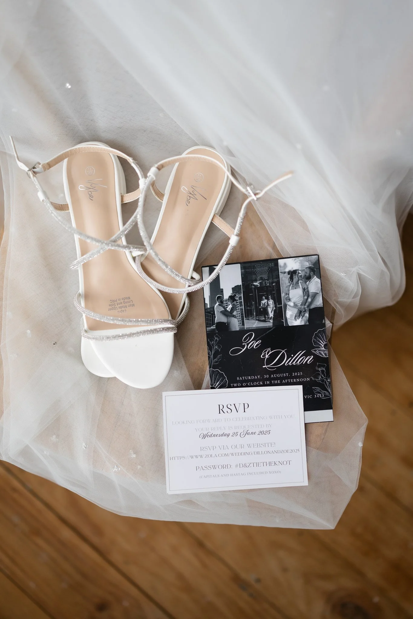 A pair of white high-heeled sandals with sparkly straps, a black and white wedding invitation featuring a couple, and an RSVP card, all laid on a white tulle fabric on a wooden floor.