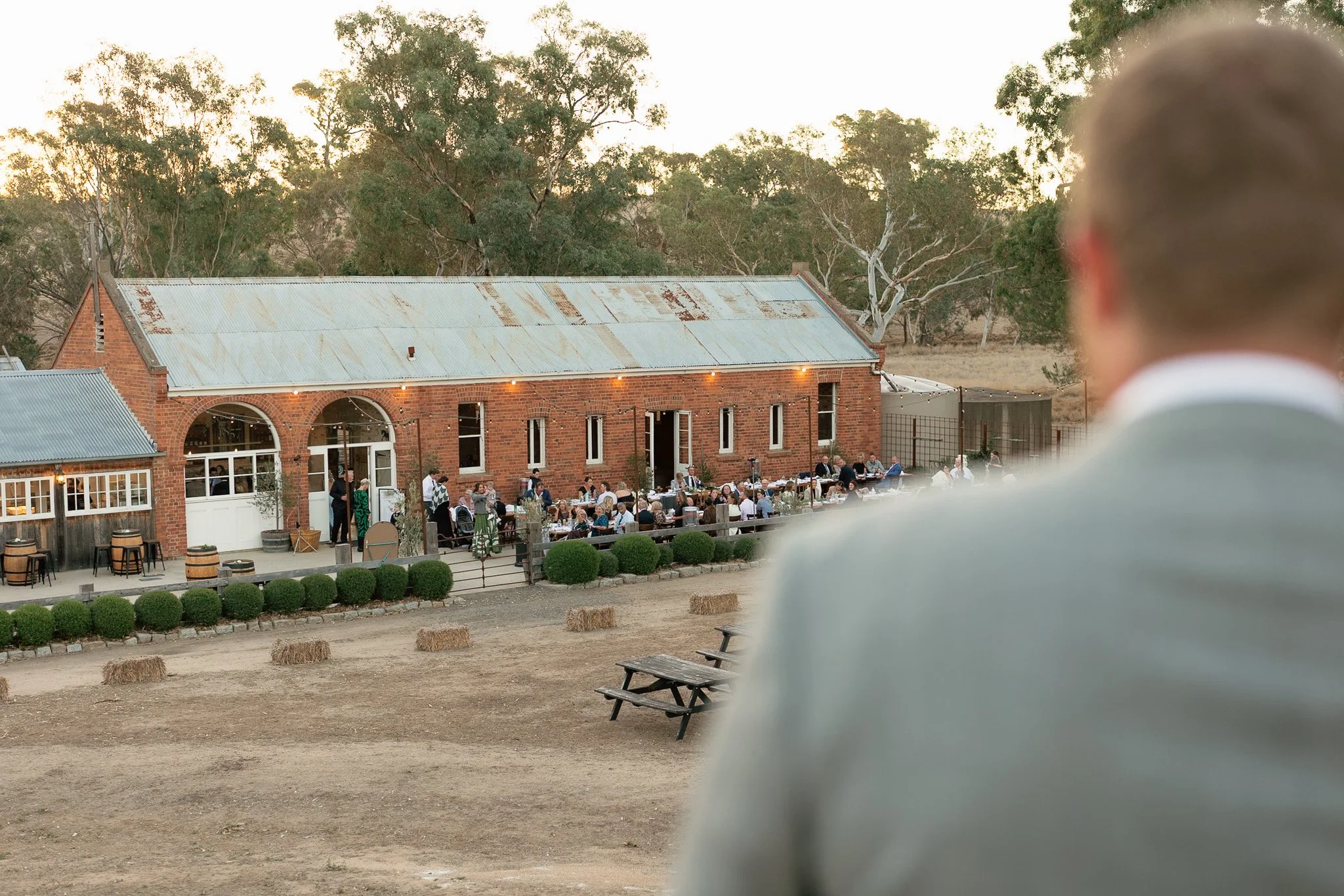 A person in the foreground looking at a rustic outdoor gathering at sunset, with a brick building and trees in the background.