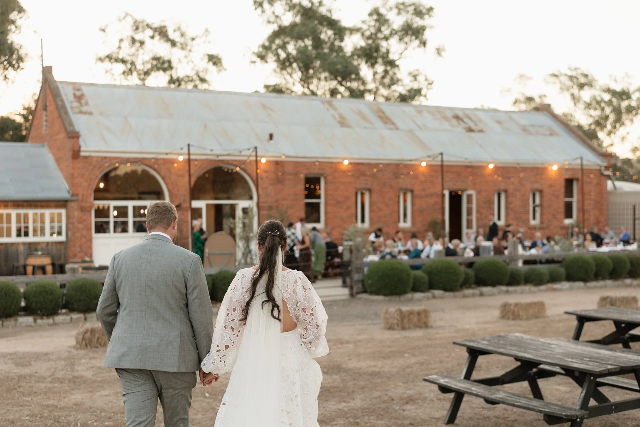 A bride and groom hold hands during their wedding ceremony outdoors, with a rustic brick barn and guests in the background.