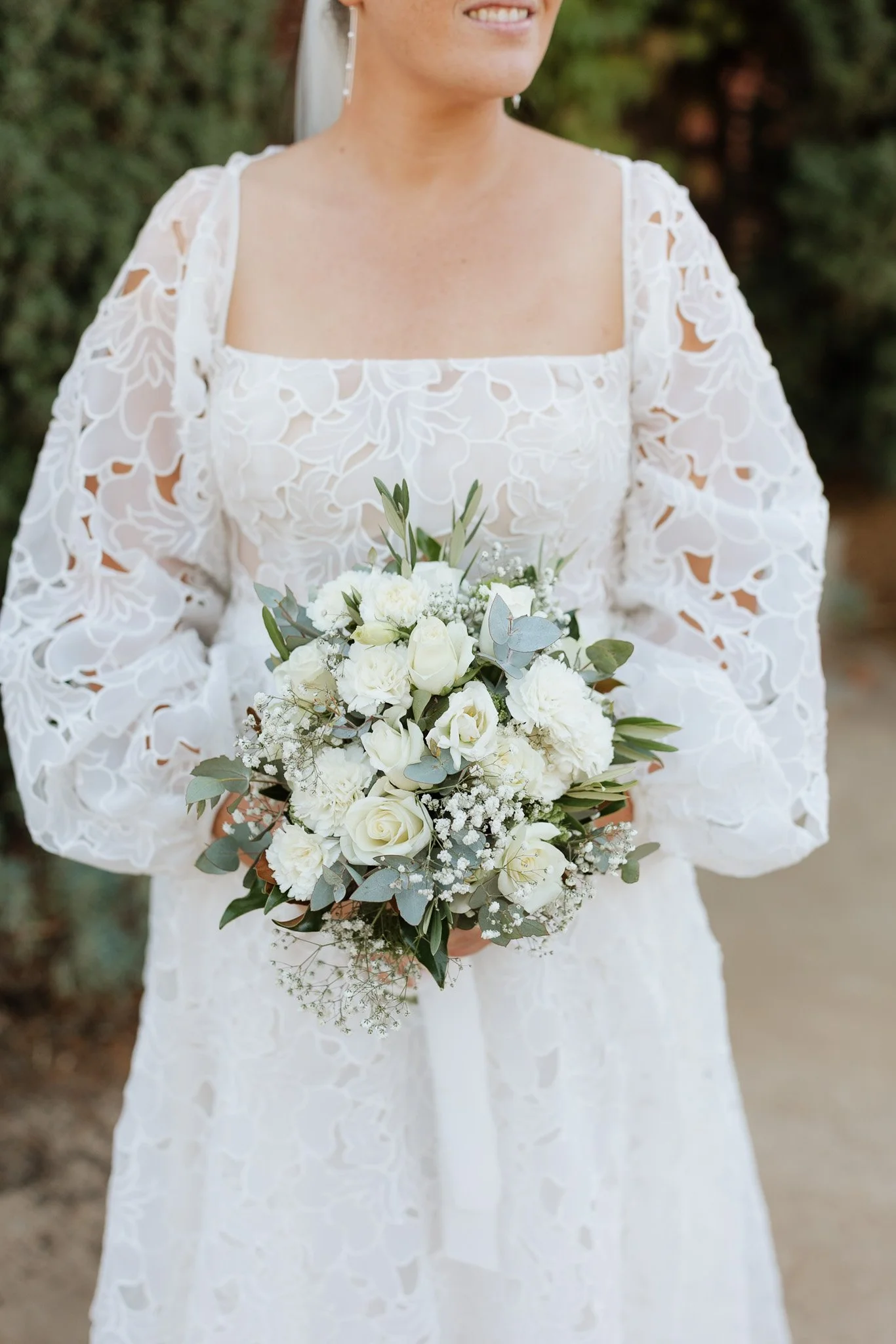 Close-up of a bride holding a bouquet of white roses and greenery, wearing a white lace wedding dress with puffed sleeves, outdoors with blurred trees in the background.