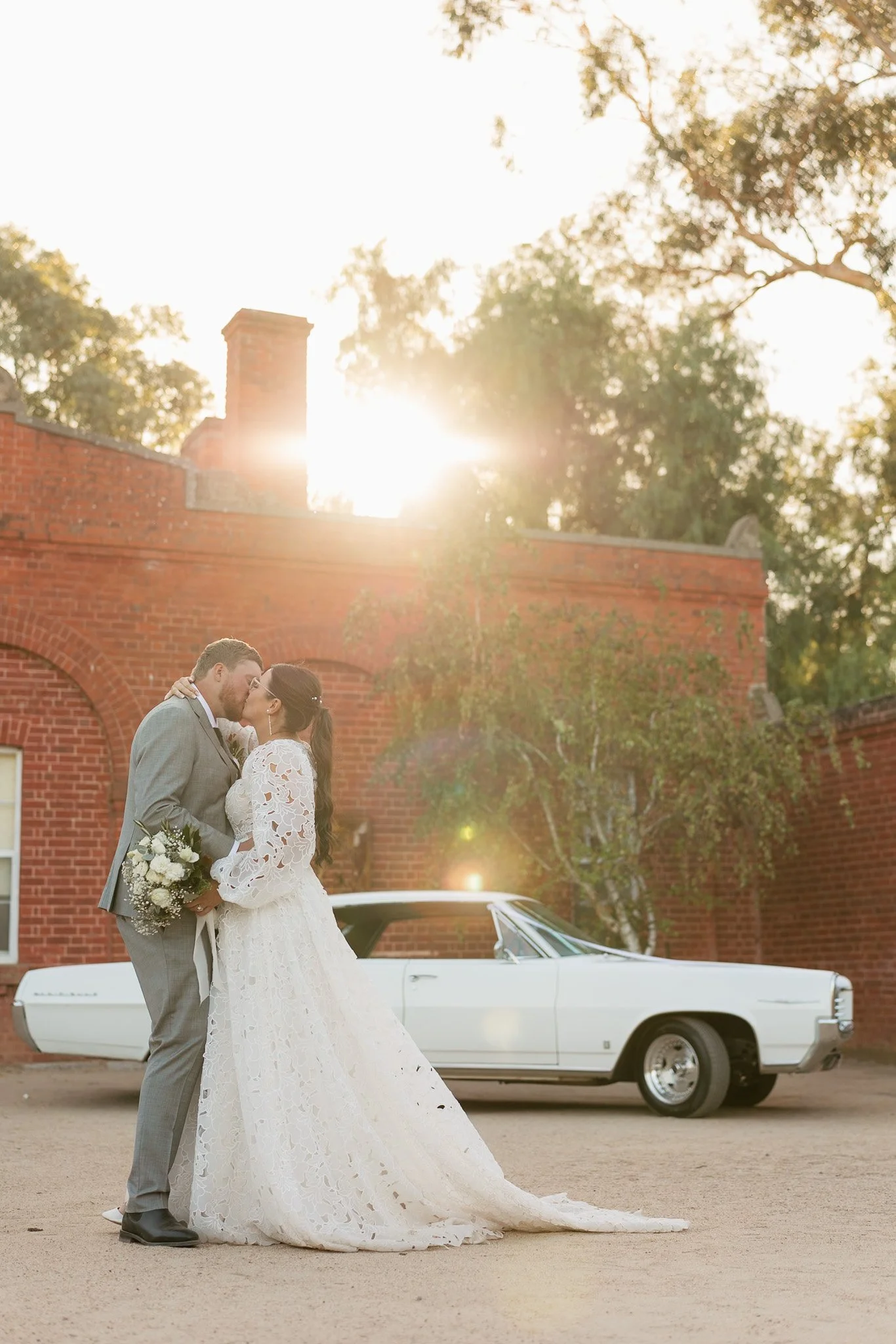 A bride and groom share a kiss outdoors during sunset, standing in front of a white vintage car and a red brick wall with green trees in the background.