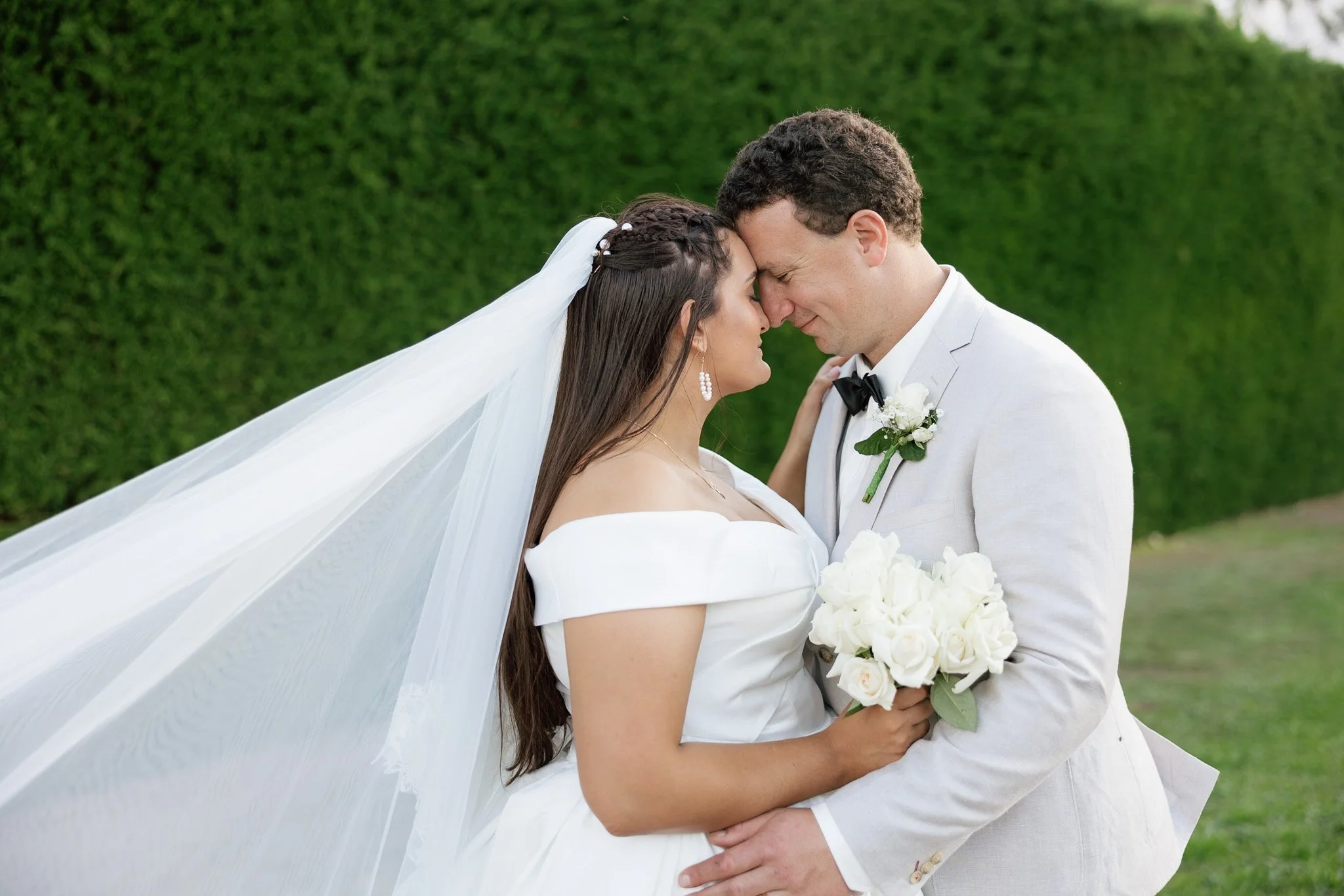 A bride and groom standing close with their foreheads touching, holding a bouquet of white flowers, outdoors with a green hedge background.