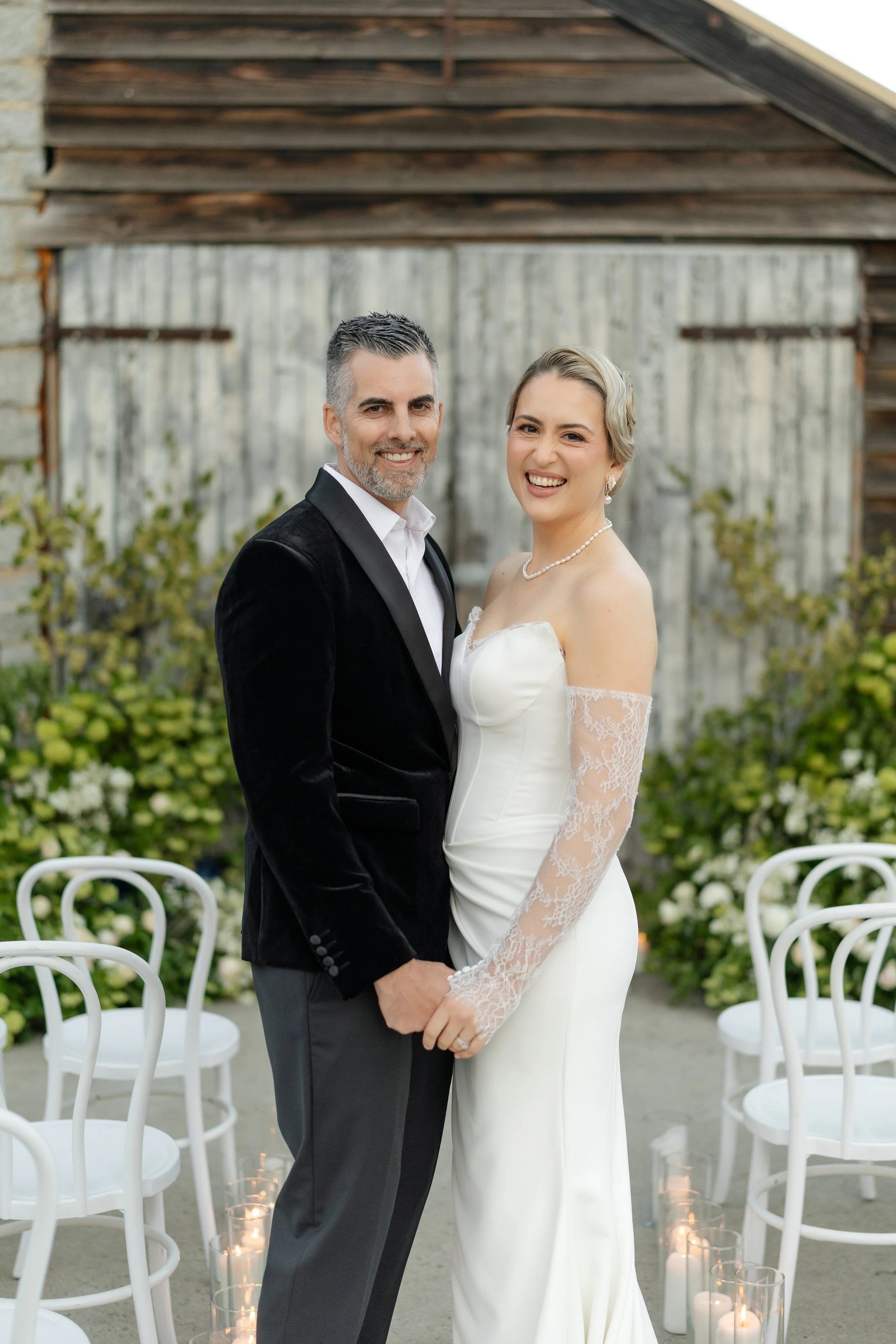 A newlywed couple standing outdoors, holding hands and smiling. The bride wears a strapless white wedding gown with lace sleeves, pearl necklace, and earrings. The groom wears a black velvet tuxedo jacket and gray pants. There are white chairs and ca