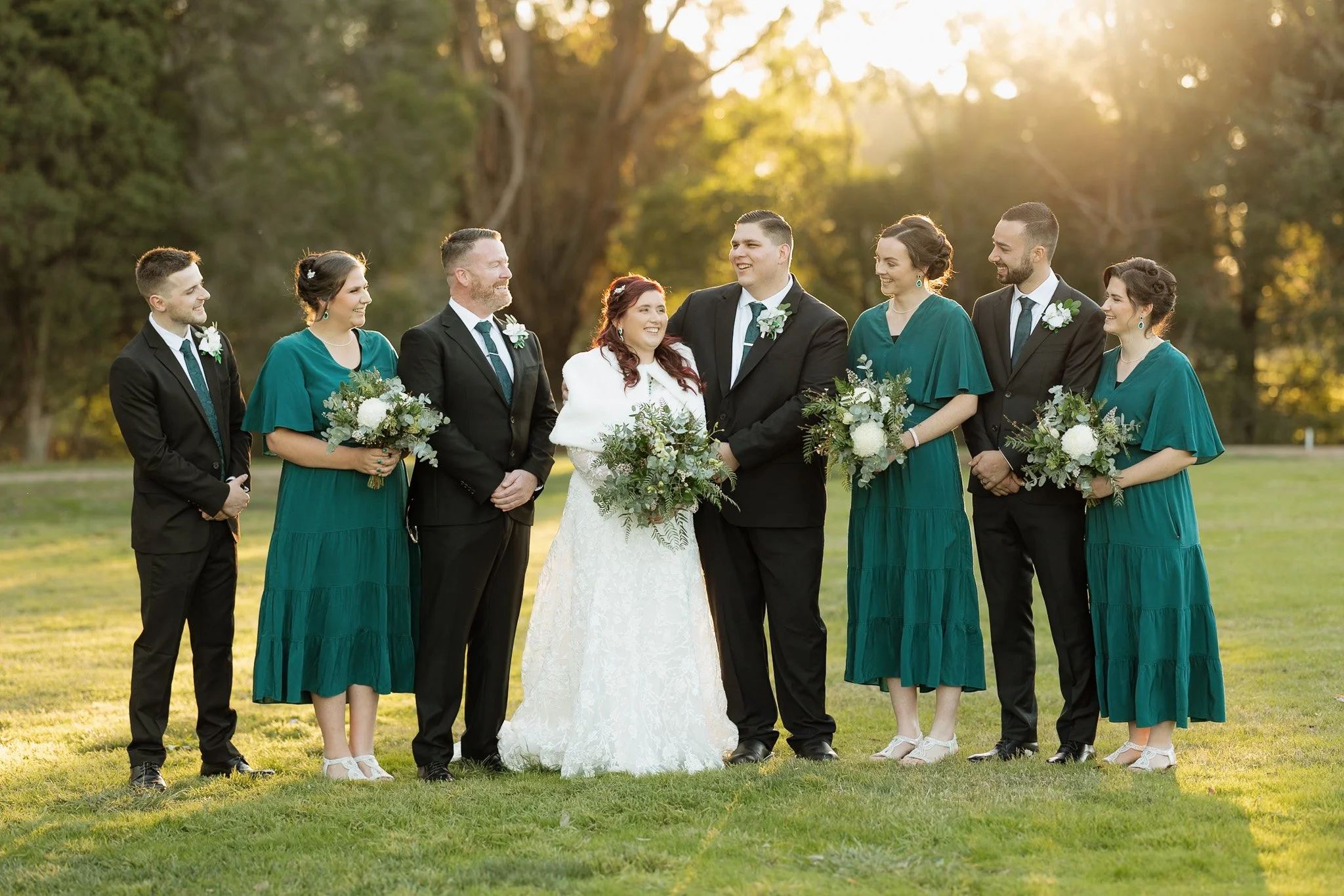 A wedding ceremony outdoors with a bride, groom, and six bridesmaids and groomsmen standing in a line on a grassy field with trees in the background, during sunset.