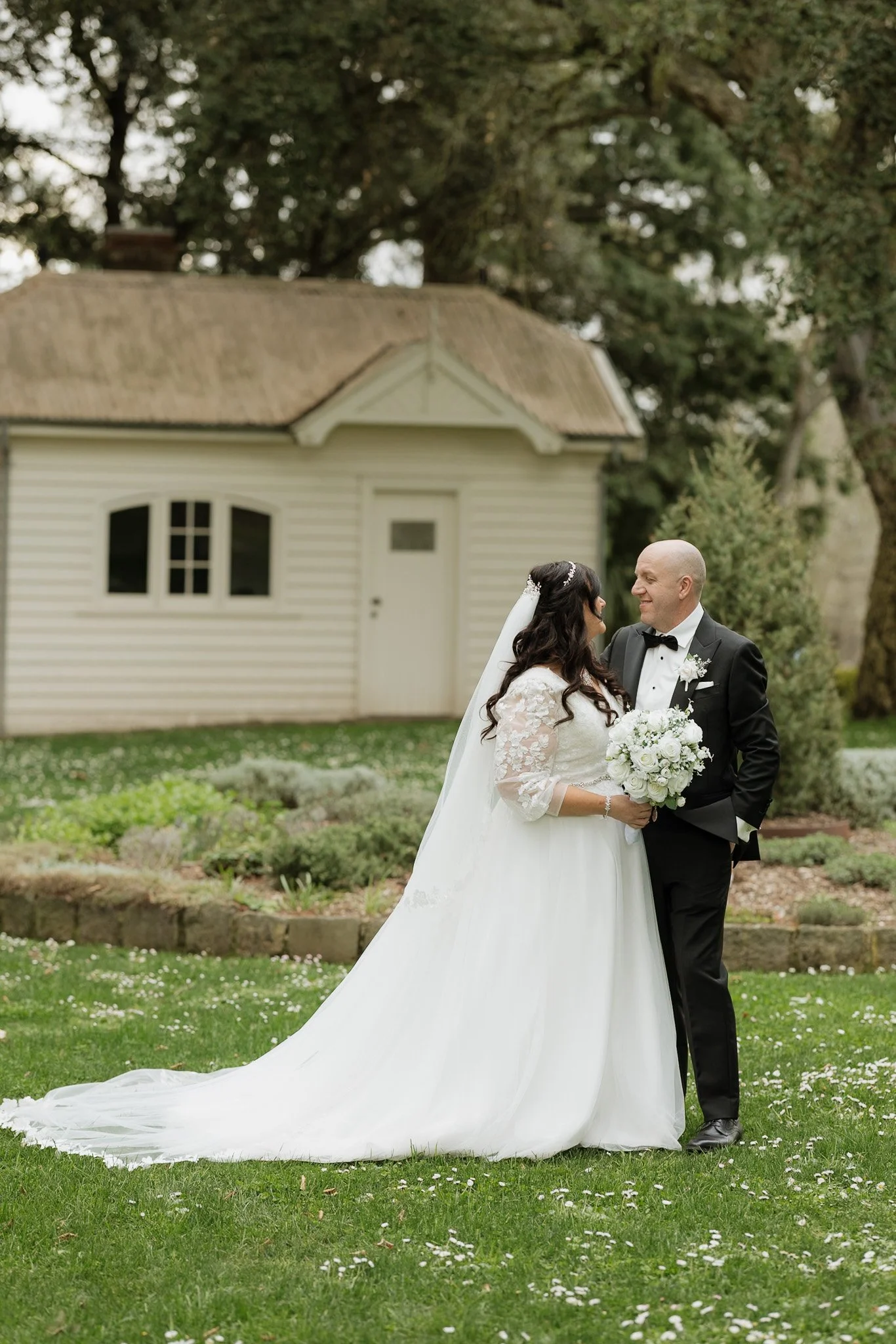 A bride and groom standing outdoors on a wedding day, with a small white house and large trees in the background. The bride is wearing a white wedding gown and holding a bouquet, while the groom is dressed in a black tuxedo.