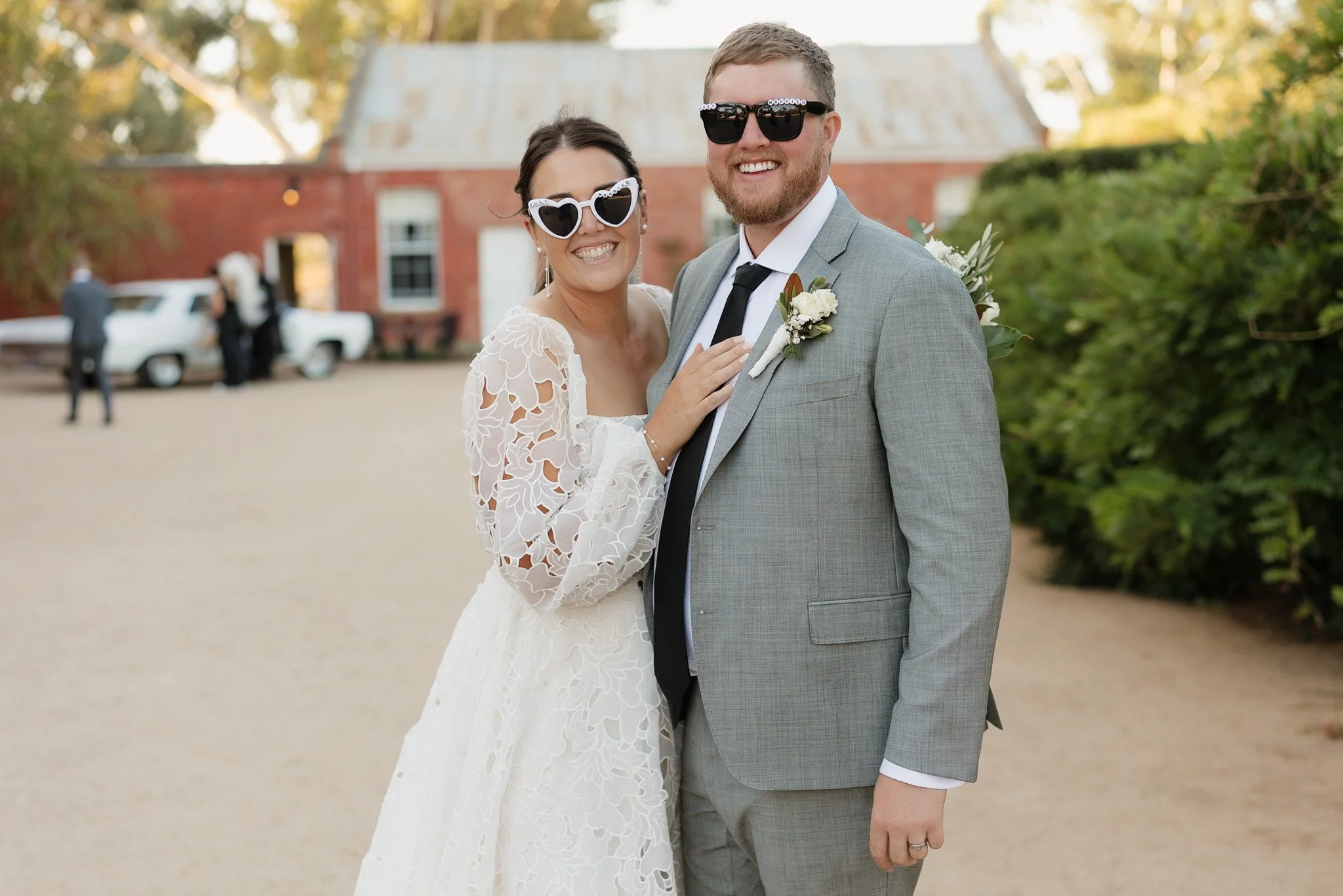 Happy couple smiling at wedding, the woman wearing white lace dress and heart-shaped sunglasses, the man in gray suit and black sunglasses, standing outdoors with wedding guests and vintage car in background.