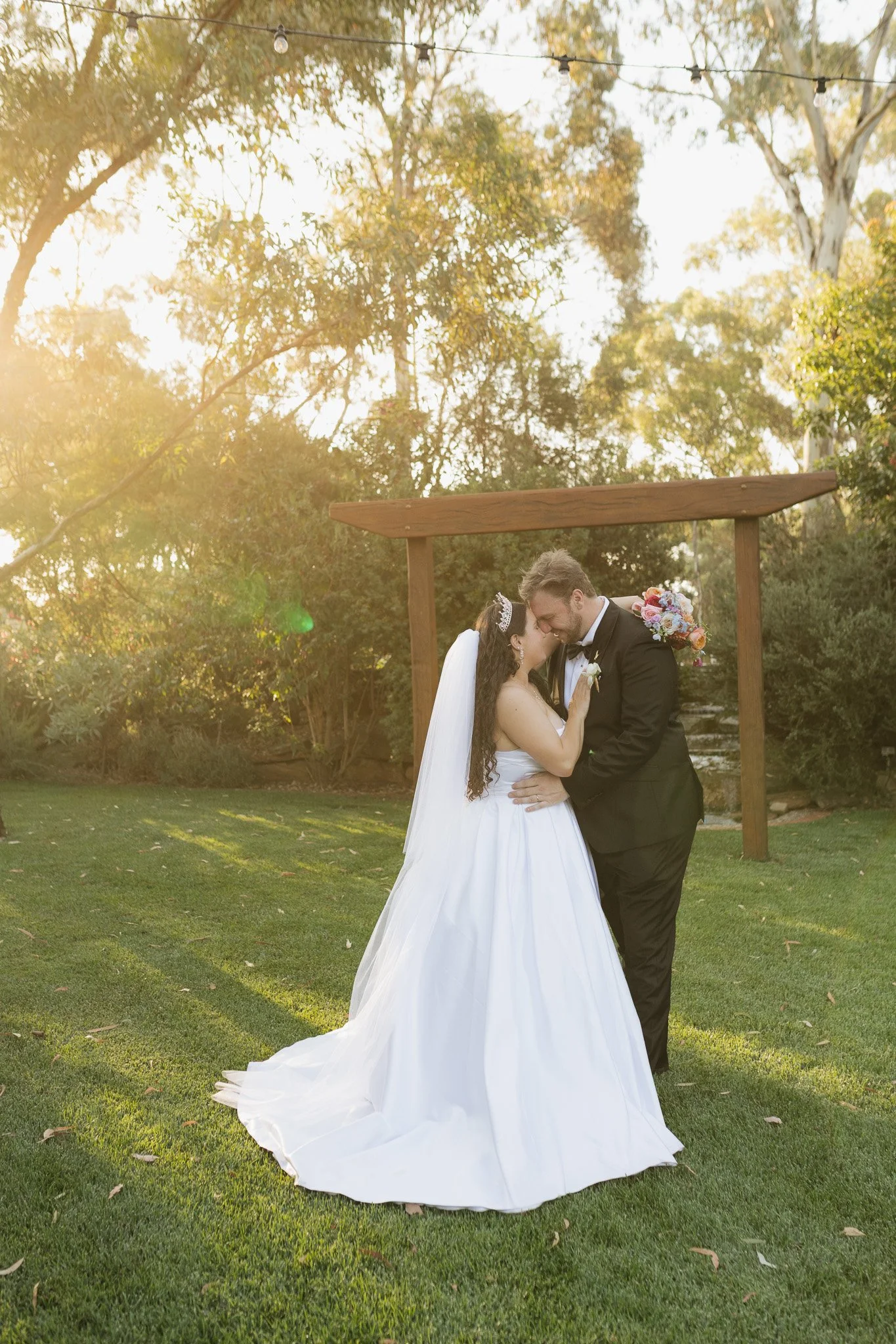 A bride and groom share a kiss during their outdoor wedding ceremony in the late afternoon, with sunlight filtering through trees in the background. The bride is wearing a white wedding gown, and the groom is in a black tuxedo. The setting is a grass