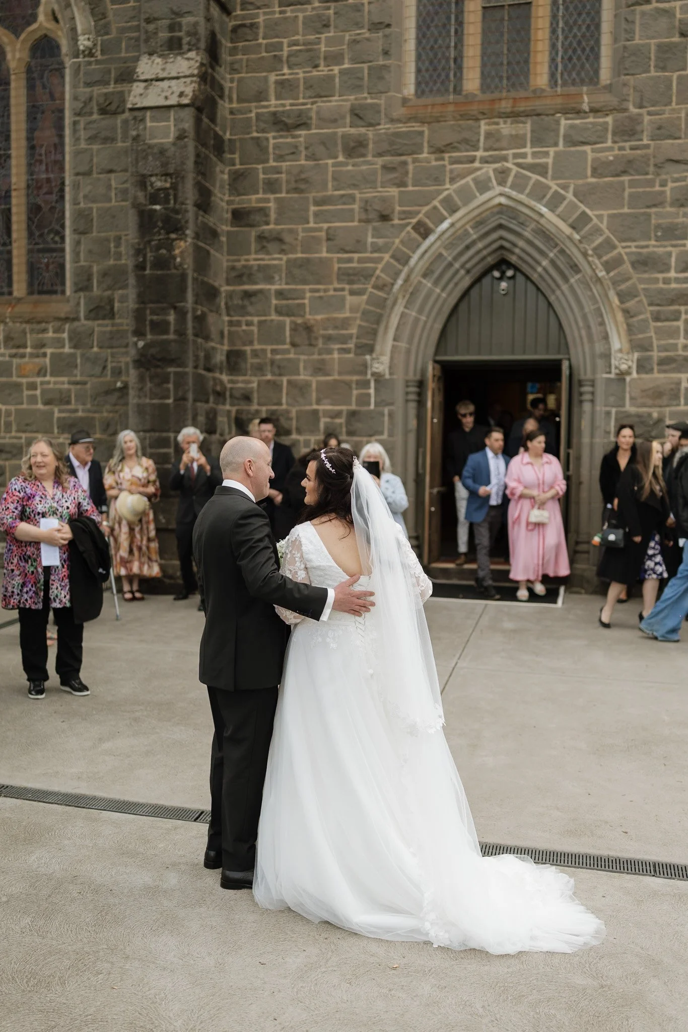 A bride and groom standing face-to-face outside a stone church, with guests gathered around them, during a wedding ceremony.