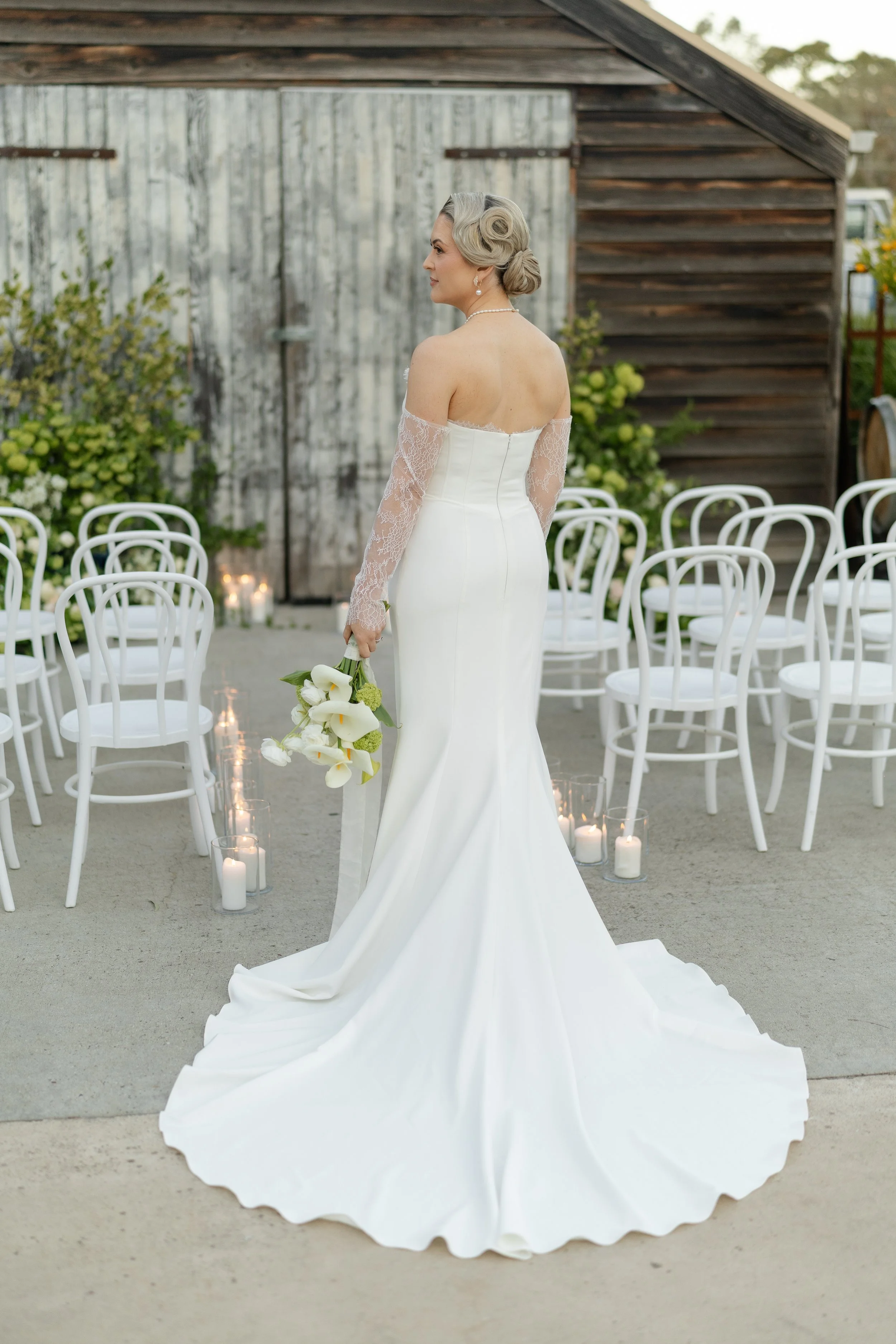 A bride in a white wedding dress with lace sleeves holding a bouquet of white calla lilies, standing outdoors with chairs and candles in the background.