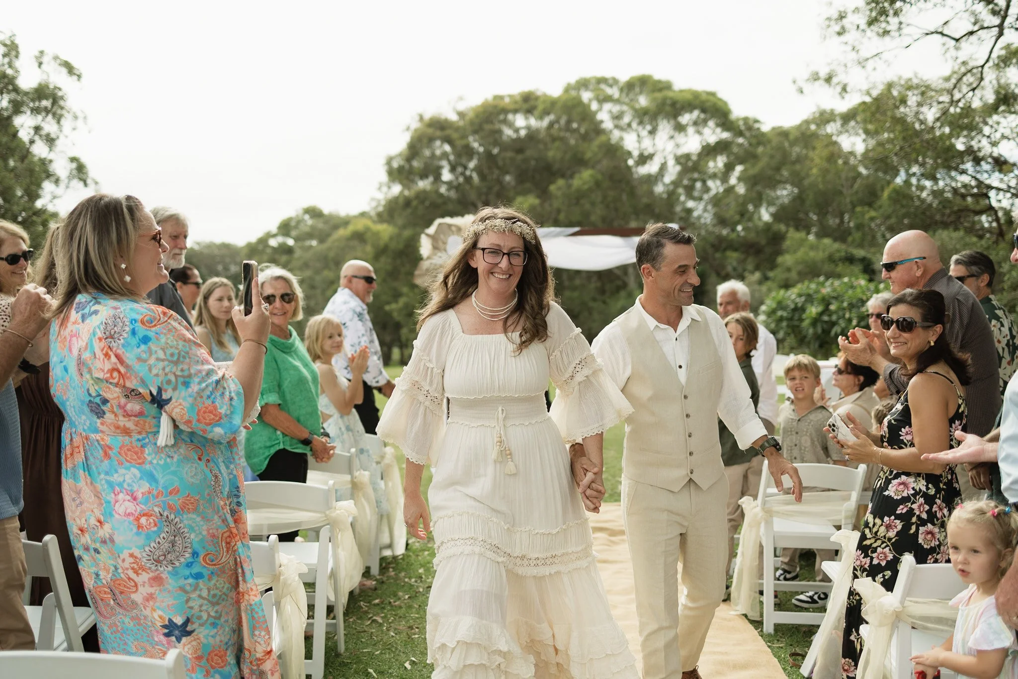 A bride and groom holding hands and smiling as they walk down the aisle at an outdoor wedding ceremony, surrounded by guests cheering and taking photos.