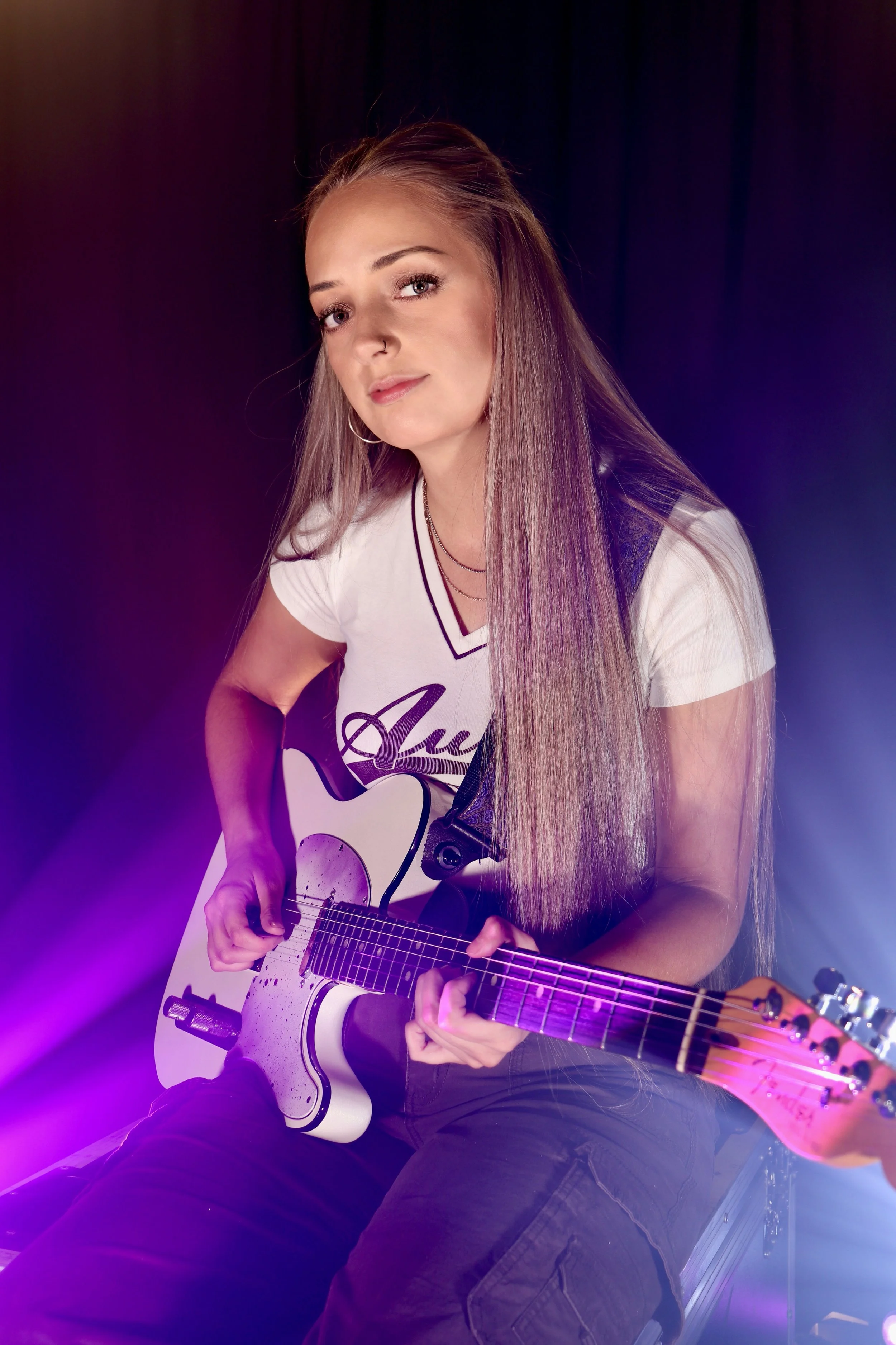 Young woman with long blonde hair playing an electric guitar on stage, wearing a white t-shirt and dark pants, with colorful stage lighting.