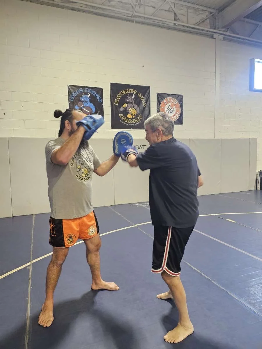 Two men practicing boxing in a gym. The man on the left is wearing a gray T-shirt, orange shorts with black and yellow logos, and blue gloves. The man on the right is wearing a black T-shirt, black shorts with red and white trim, and blue gloves, and he is throwing a punch while the man on the left blocks with his glove. There are three gym banners on the wall behind them, featuring a bull, a bear, and a fist, with the text "Haverrick Wake Academy," 
