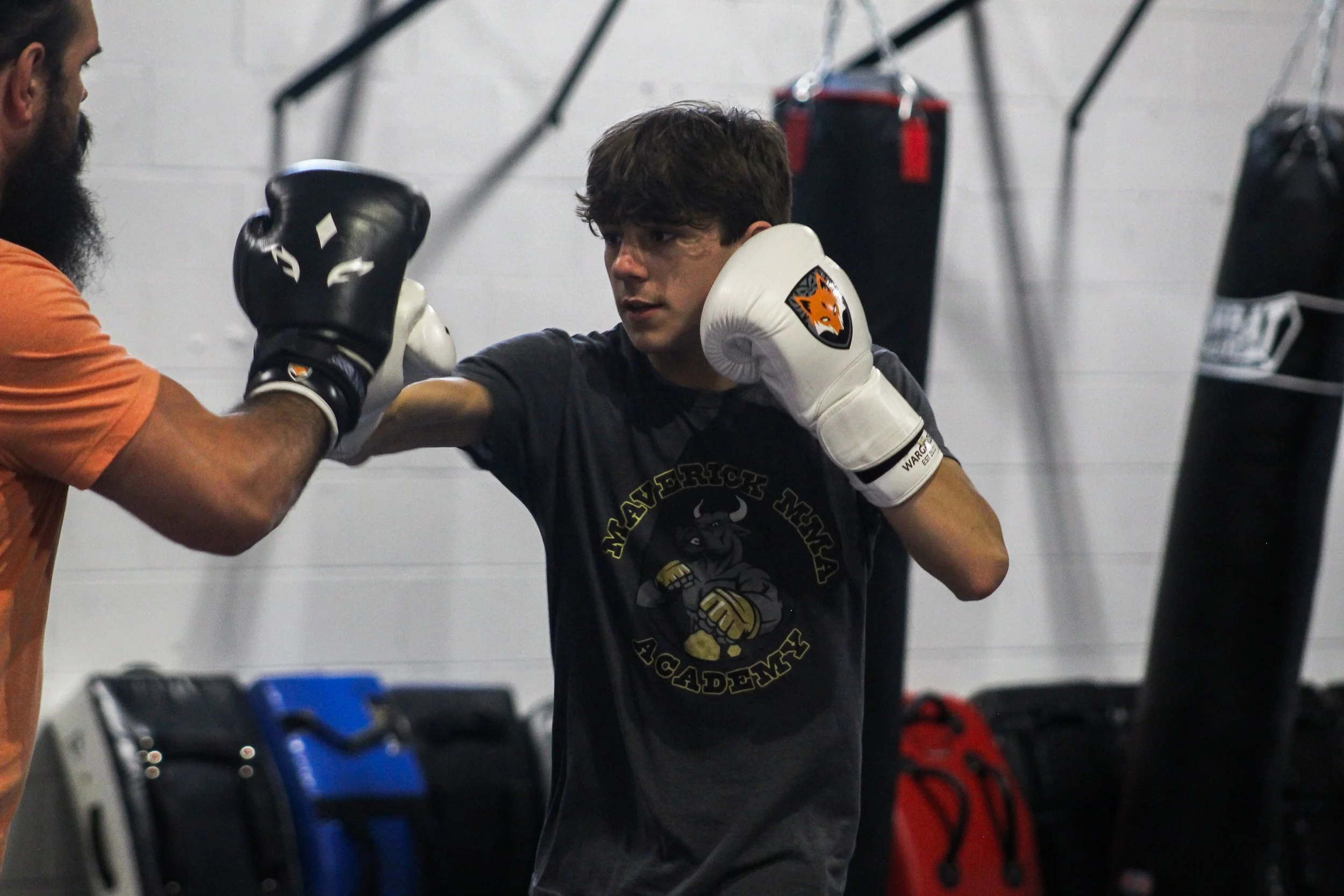 A young male boxer practicing punches with a trainer in a gym, while wearing boxing gloves and sportswear.
